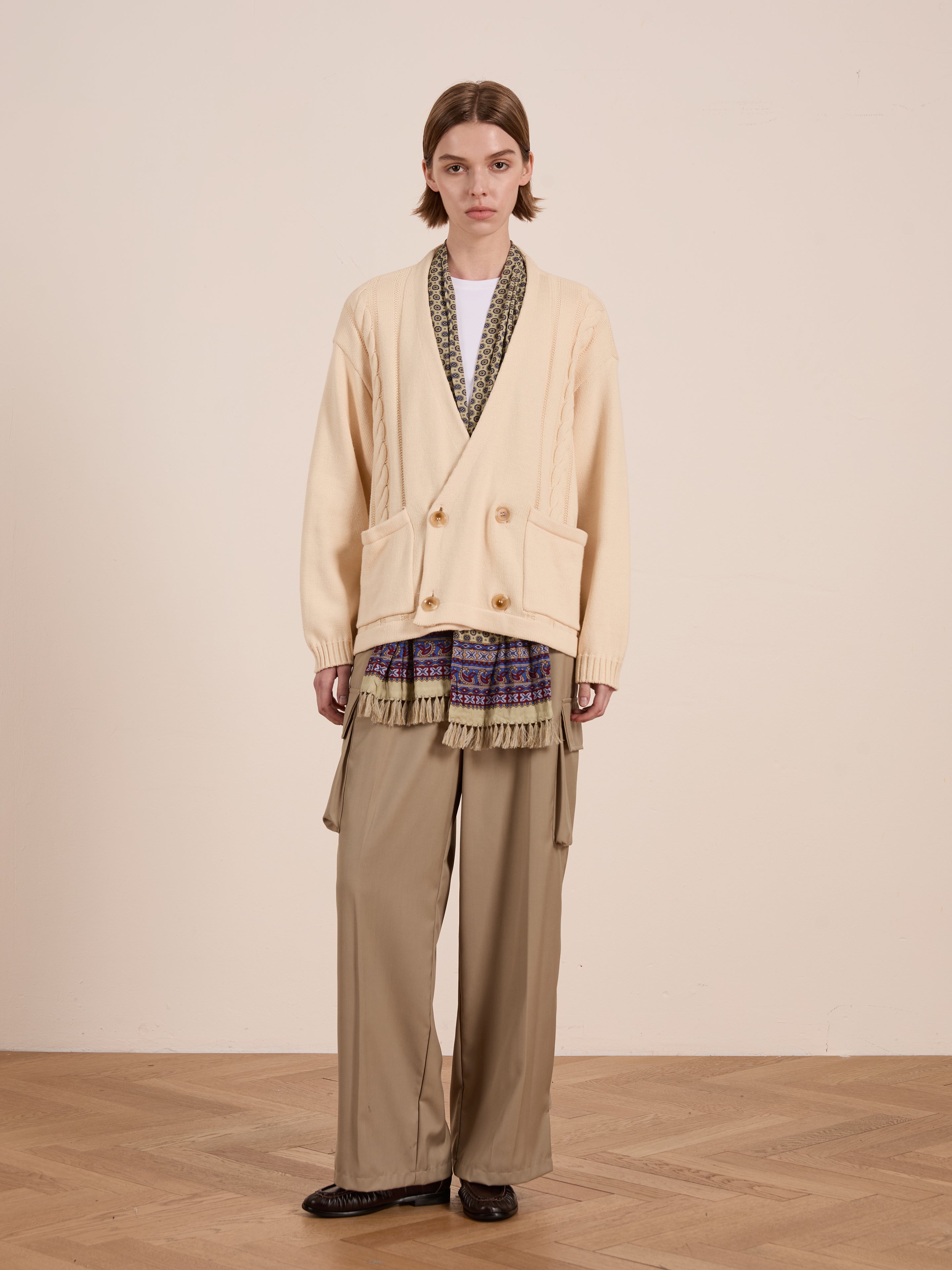 A person stands on a wooden floor against a light beige background, wearing the cream Double Breasted Knit Cardigan by FOUND, in a relaxed fit. A patterned scarf, layered skirt, unisex wide-leg tan trousers, and dark shoes complete the look.