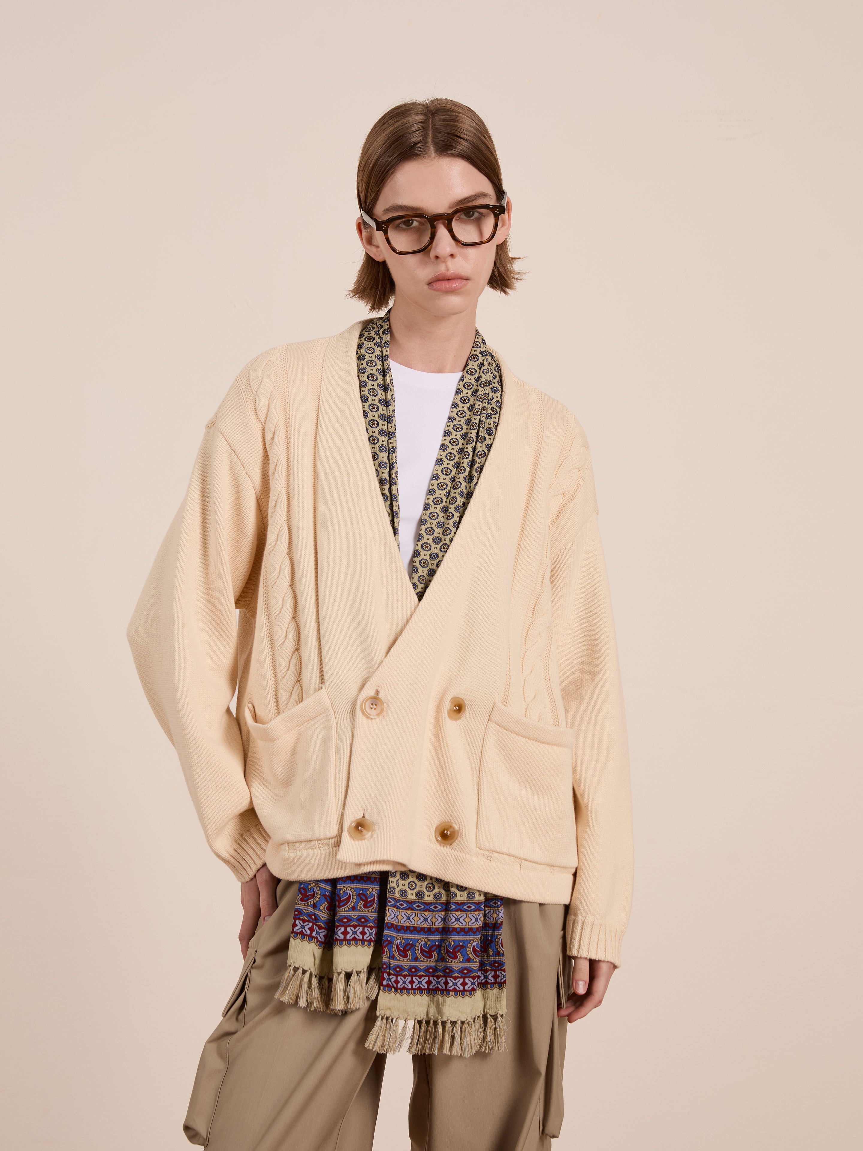 A person with shoulder-length hair and glasses stands against a plain background, wearing a relaxed fit beige Double Breasted Knit Cardigan by FOUND, paired with a patterned scarf and khaki pants. The unisex ensemble exudes effortless chic.