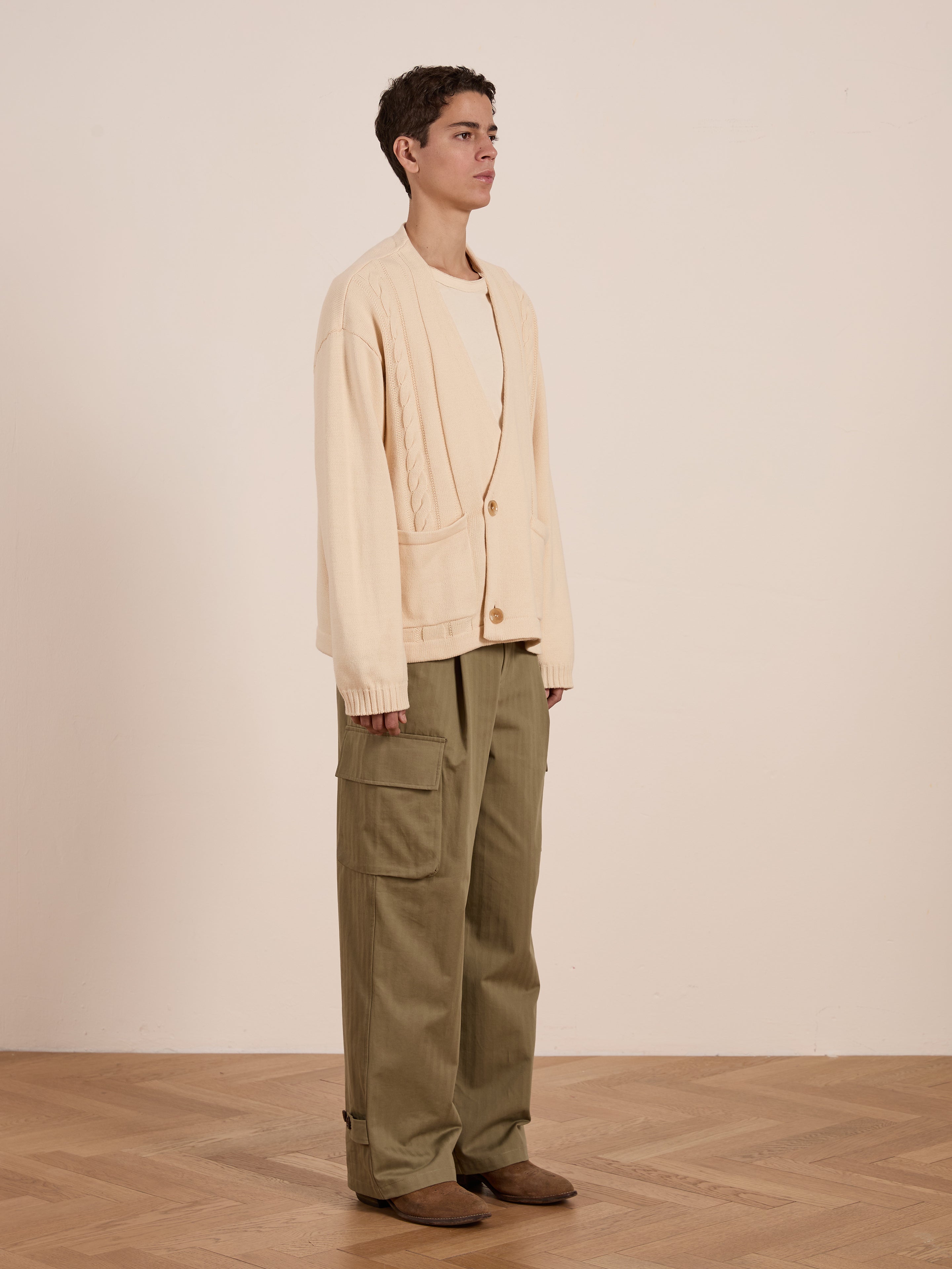 Standing indoors on a wooden floor against a plain wall, the person wears FOUND's beige Double Breasted Knit Cardigan over a shirt. Olive cargo pants and brown shoes round off the versatile look.