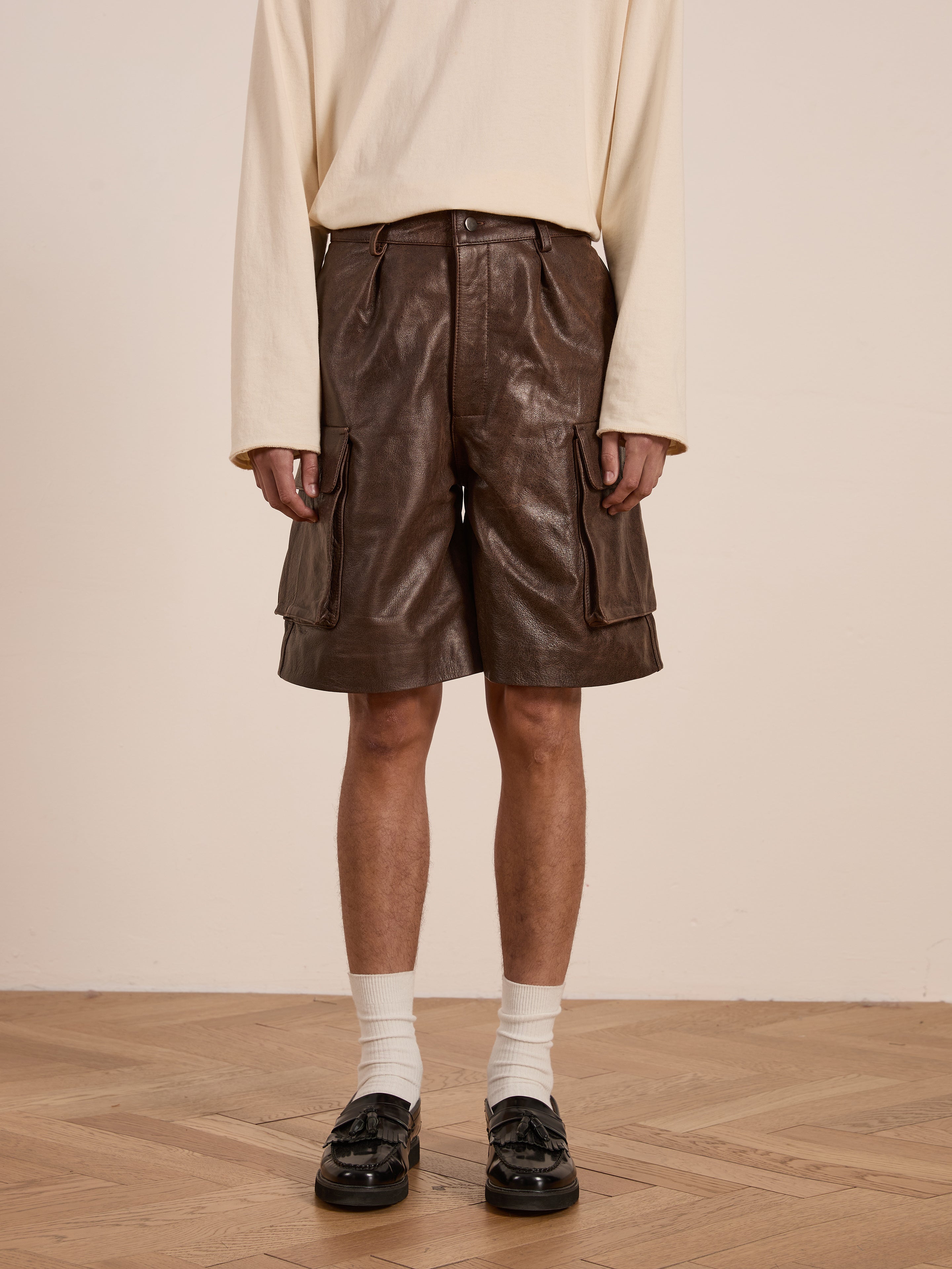 Someone wearing a long-sleeve beige shirt and FOUND's Dusk Distressed Genuine Leather Cargo Shorts stands on a wooden floor in white socks and black loafers. The minimalist room reflects their utilitarian style.