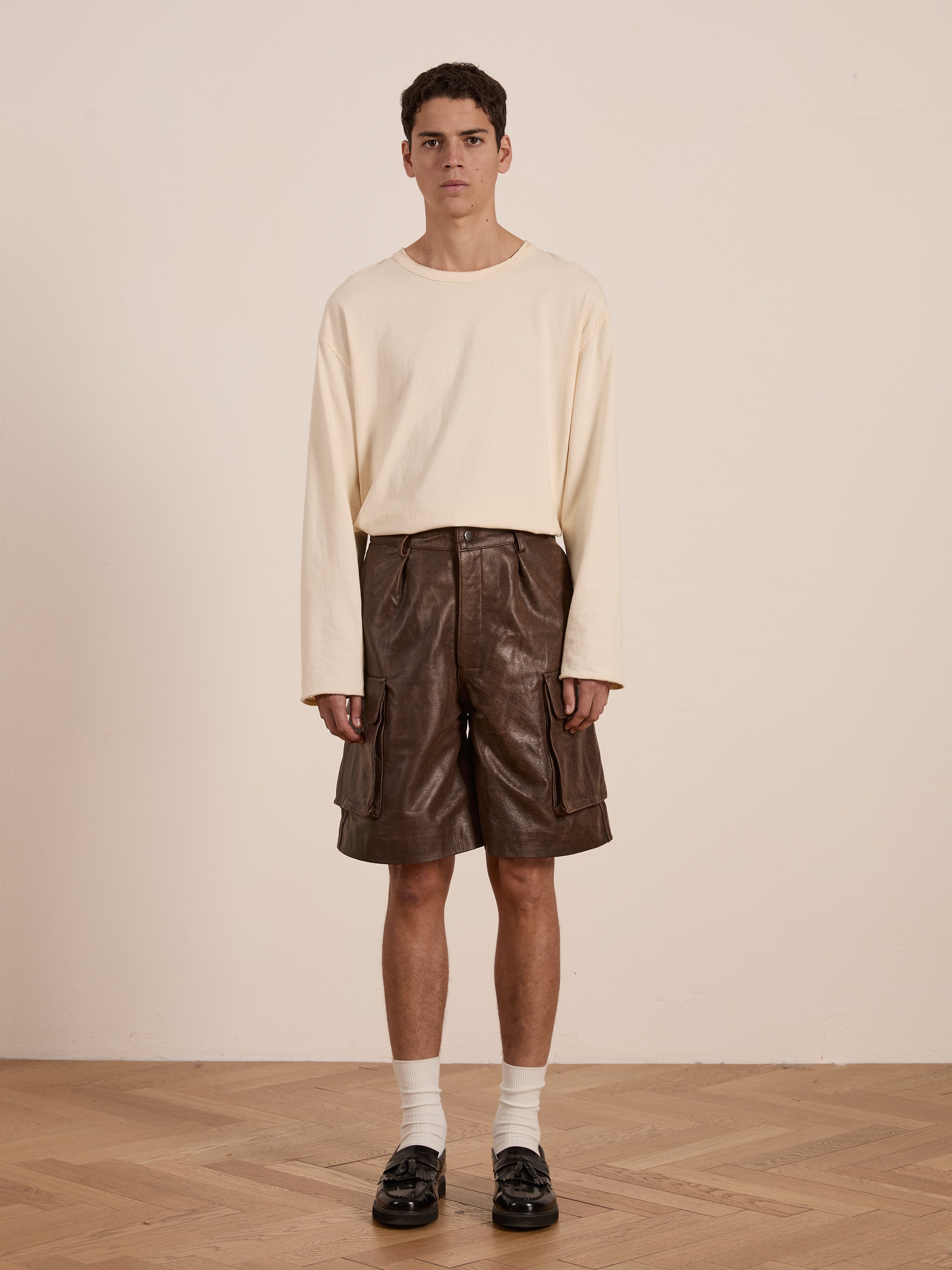 Indoors on a wooden floor, a person showcases their utilitarian roots in a cream long-sleeve shirt paired with FOUND's Dusk Distressed Genuine Leather Cargo Shorts, complemented by white socks and black shoes.