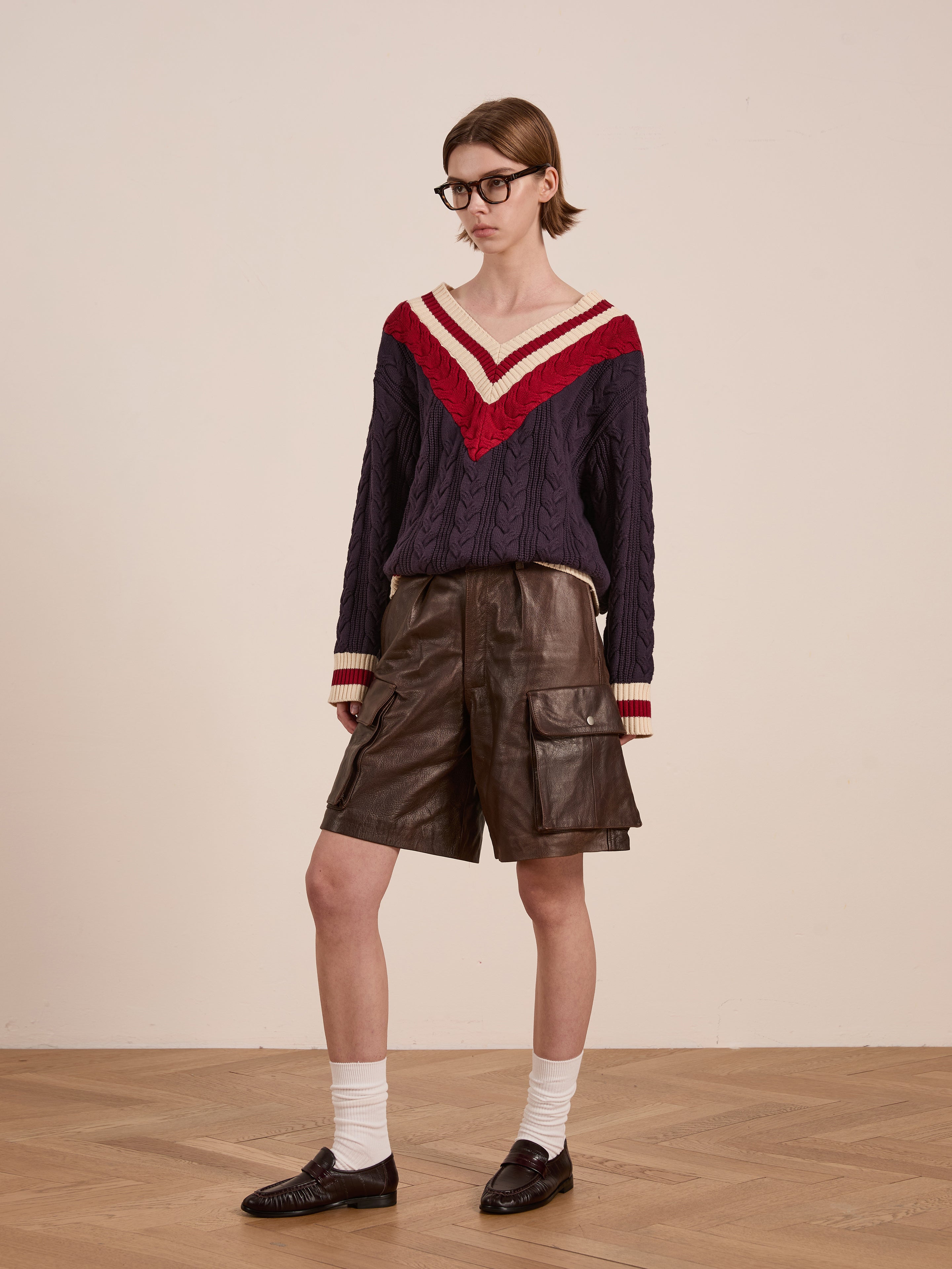 A person in glasses and a navy and red V-neck sweater stands confidently on a wooden floor, wearing white socks and brown loafers. Their FOUND Dusk Distressed Genuine Leather Cargo Shorts subtly nod to utilitarian roots against the plain backdrop.