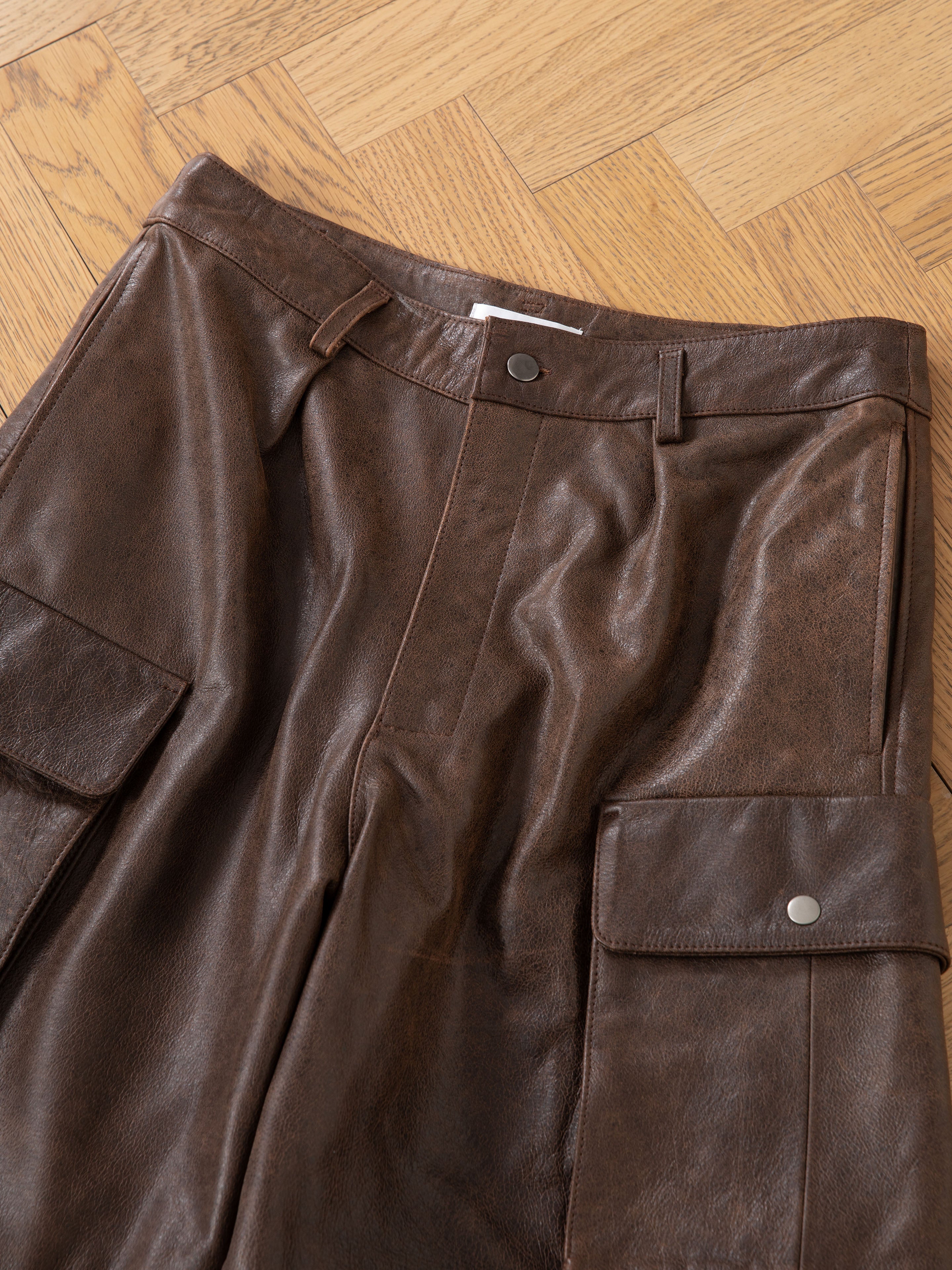 FOUND's Dusk Distressed Genuine Leather Cargo Shorts, with utilitarian roots, featuring two side pockets and belt loops, laid flat on a wooden herringbone floor.