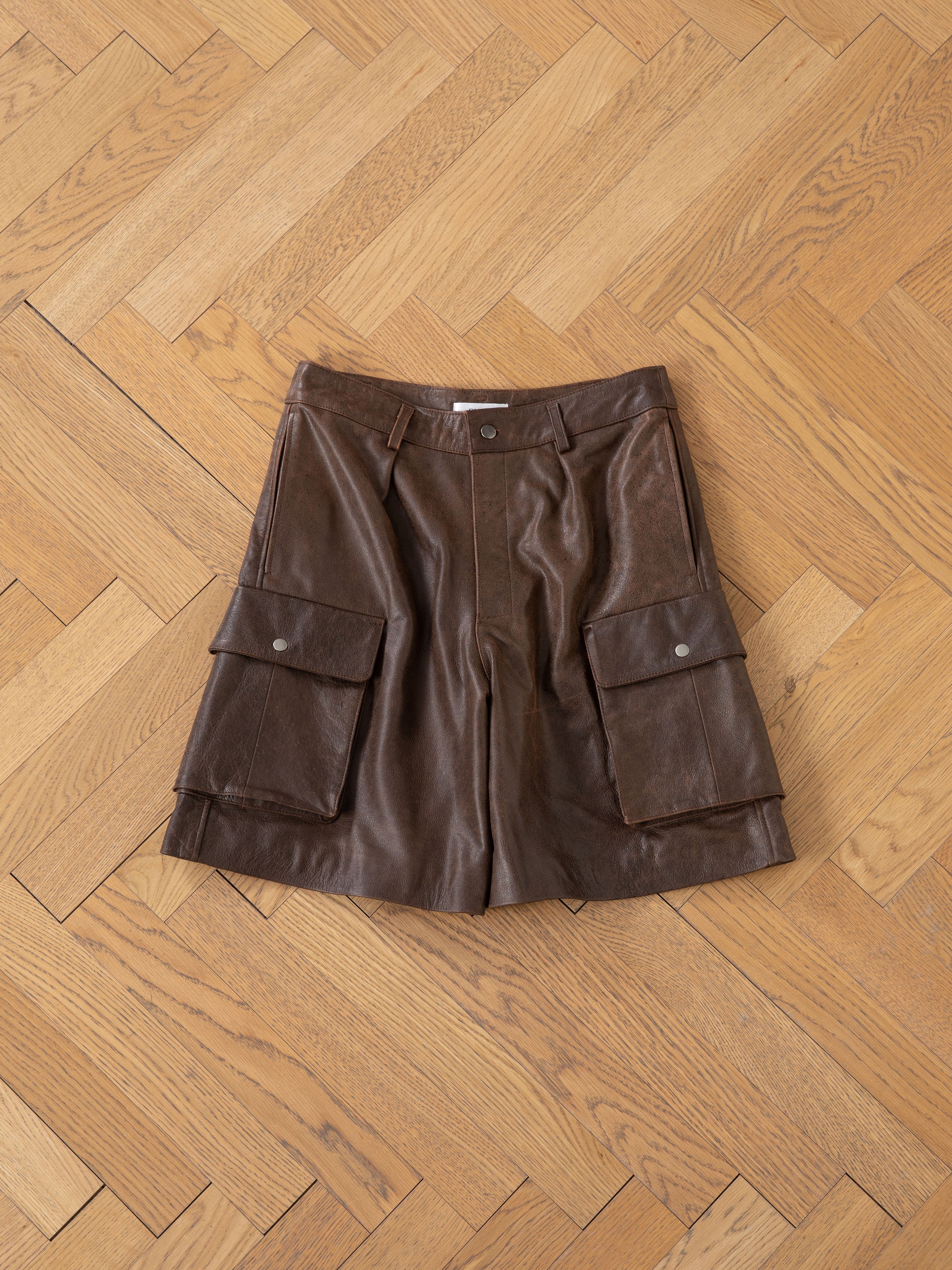 The Dusk Distressed Genuine Leather Cargo Shorts by FOUND, made from ethically sourced materials, feature large front pockets and are laid flat on a wooden parquet floor.