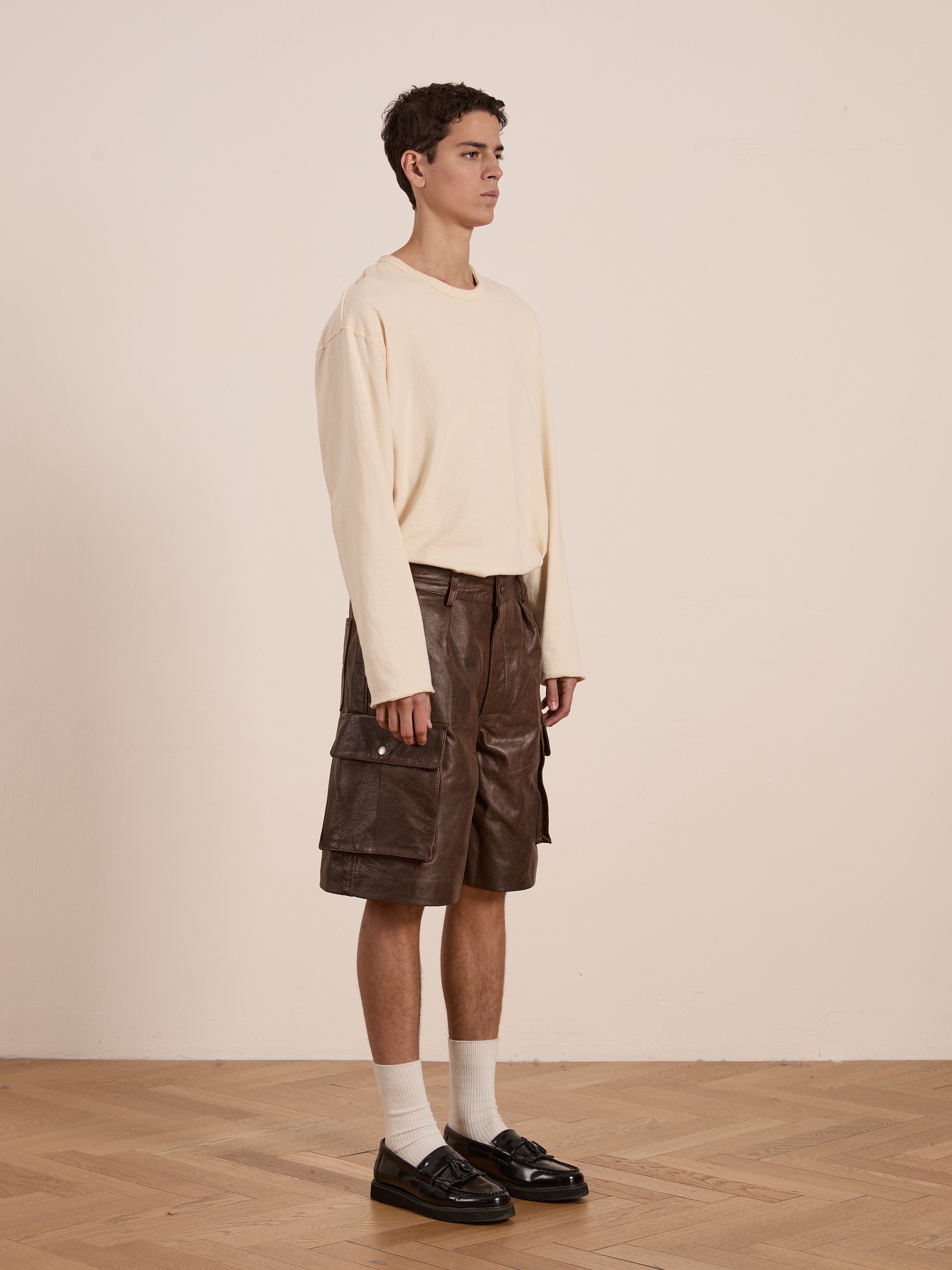 A person stands on a wooden floor, wearing a cream long-sleeve shirt and black loafers. Their white socks pair with FOUND's Dusk Distressed Genuine Leather Cargo Shorts, echoing utilitarian roots against a plain backdrop.