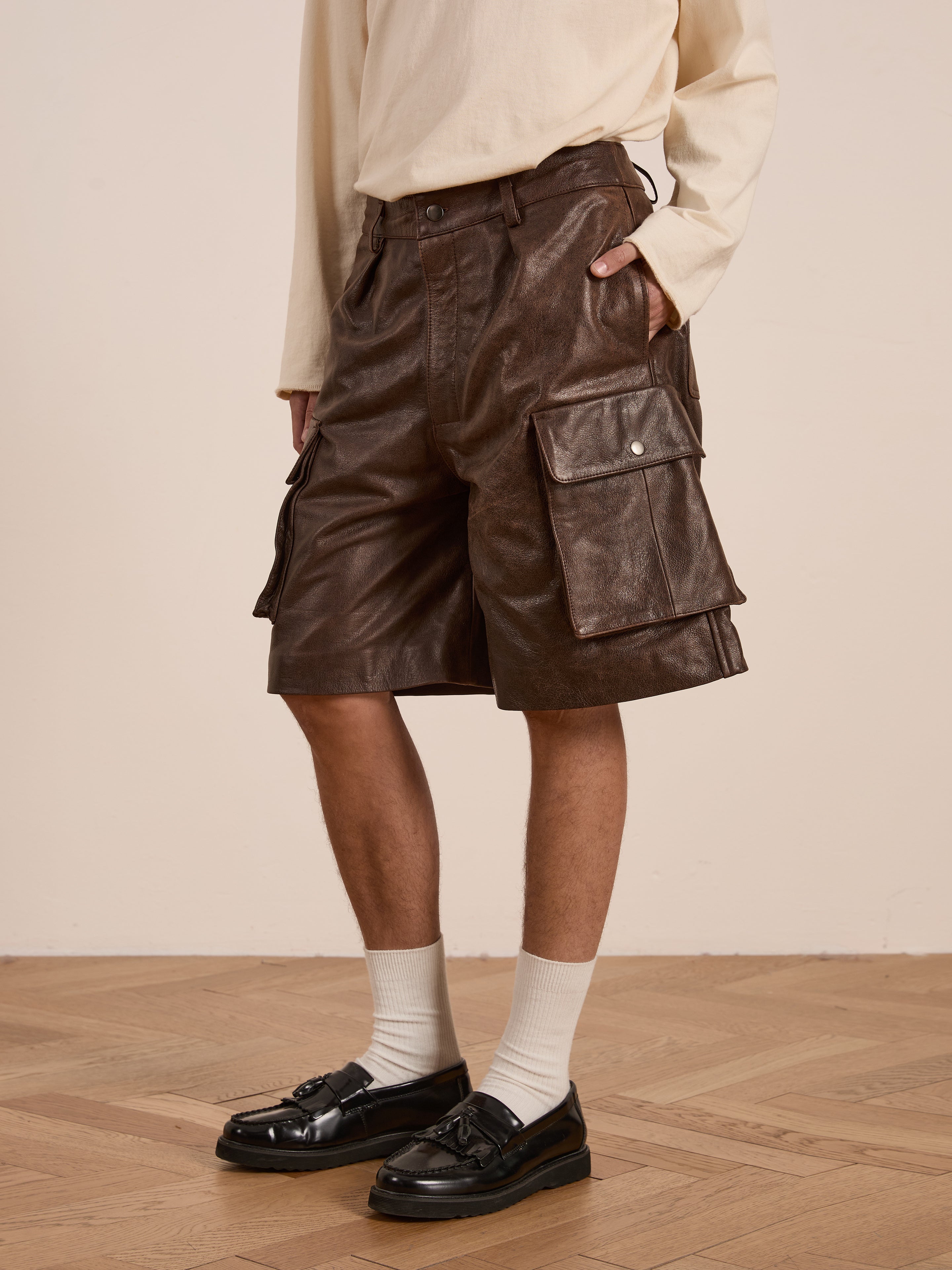 A person stands on a wooden floor, wearing FOUND's Dusk Distressed Genuine Leather Cargo Shorts in brown and a cream long-sleeve shirt. White socks and black loafers complete this look, blending utilitarian roots with an eye for detail and durability.