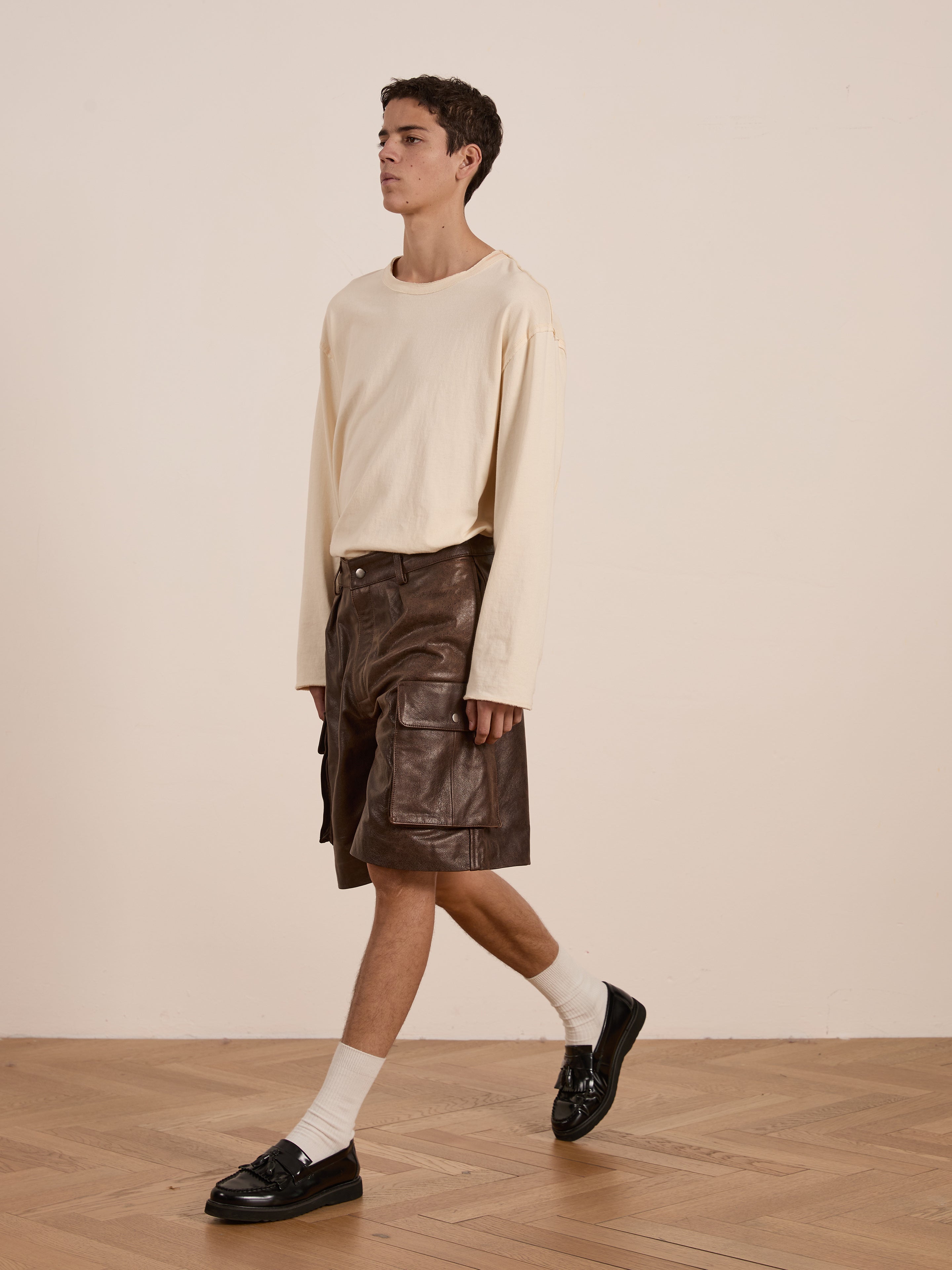 A person walks on a wooden floor, wearing a cream long-sleeve shirt and black loafers. They don FOUND's Dusk Distressed Genuine Leather Cargo Shorts, crafted from ethically sourced materials with white socks, blending utilitarian roots and contemporary style.