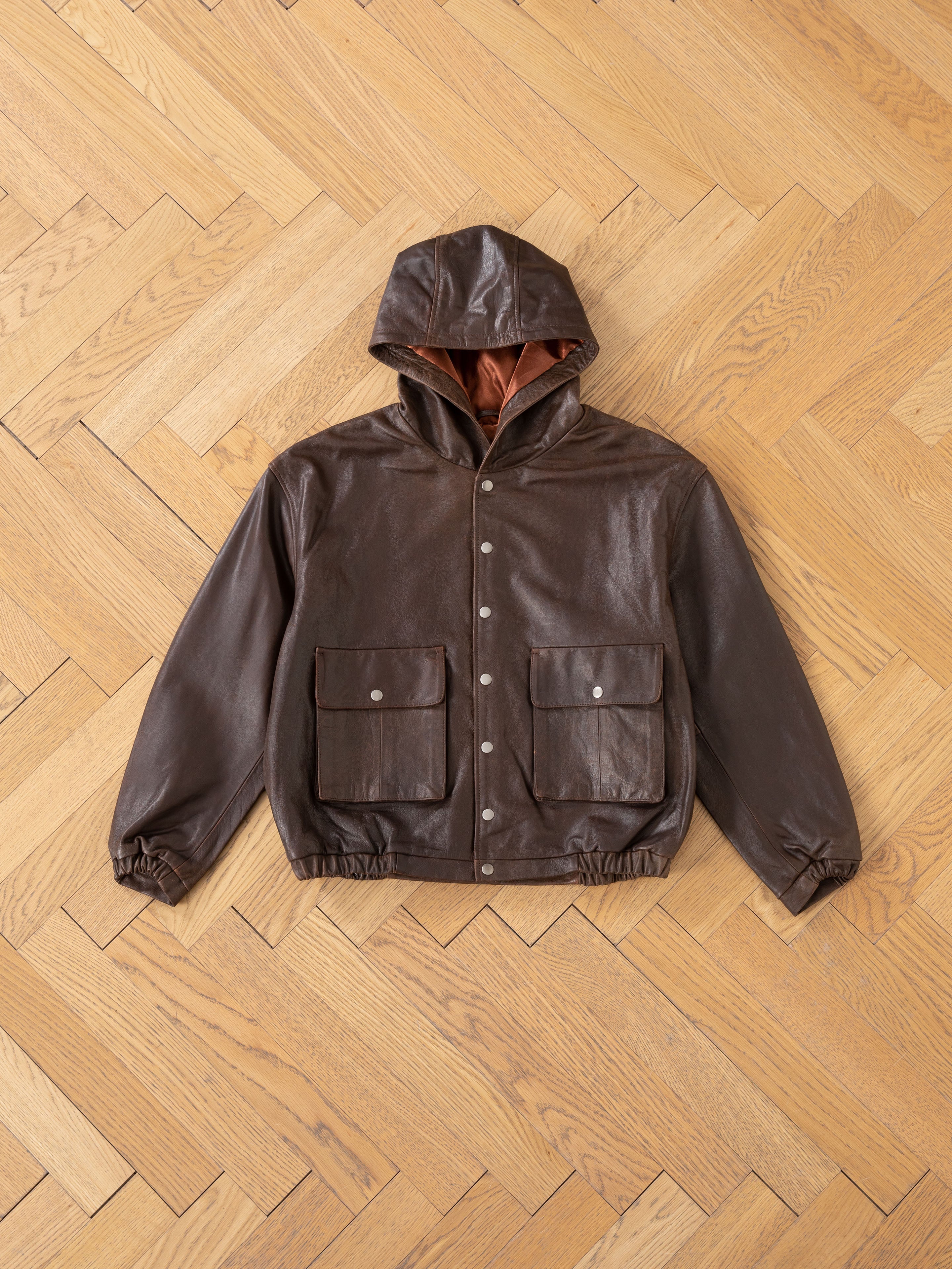 The Dusk Distressed Genuine Leather Hoodie by FOUND, made from ethically sourced materials, features a brown cowhide design with two front pockets, showcased on a wooden herringbone floor.