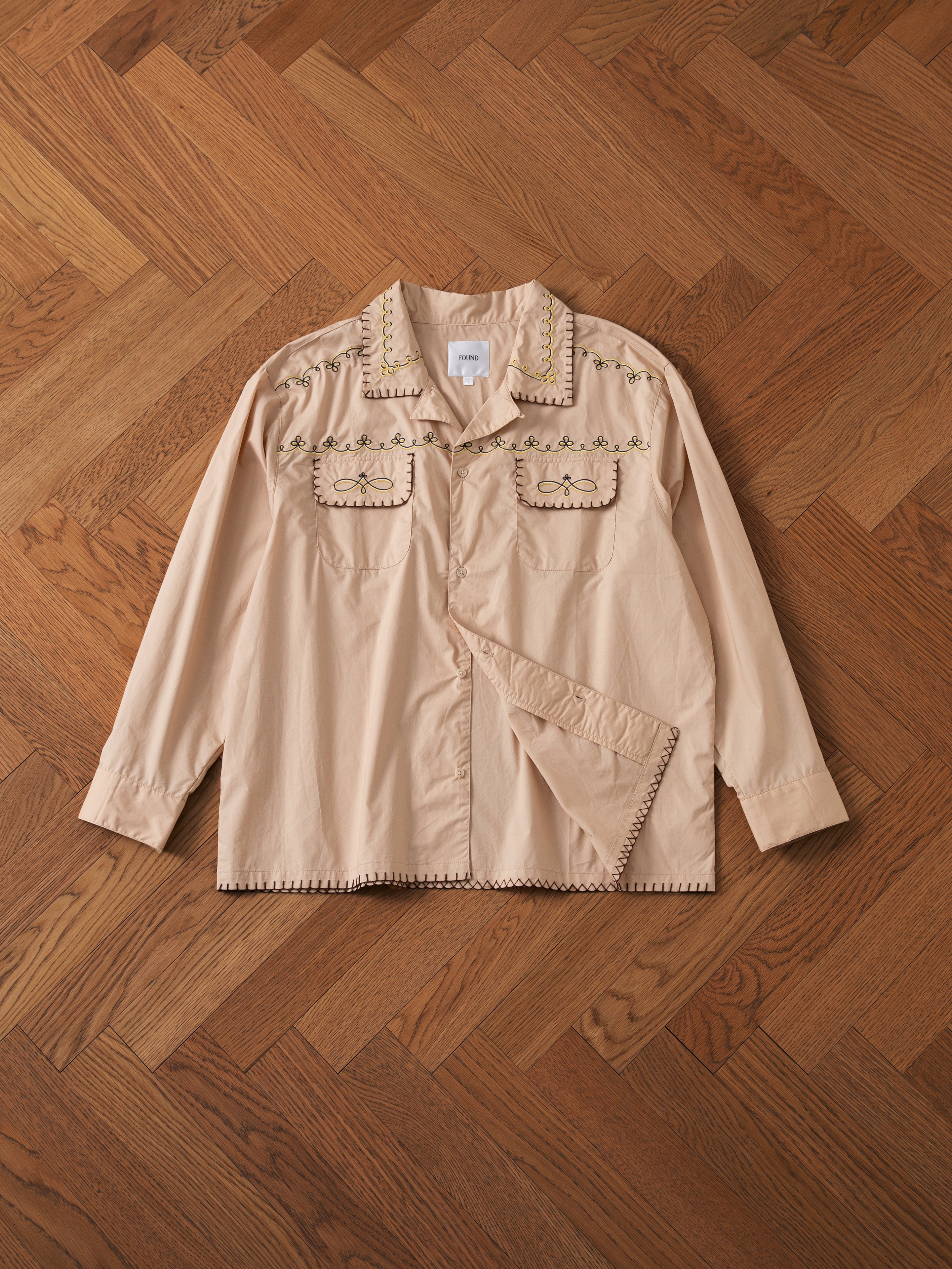 An Embroidered Western Button Up by FOUND in beige, featuring long sleeves and elegant embroidered details on the pockets and collar, displayed flat on a wooden herringbone-patterned floor.