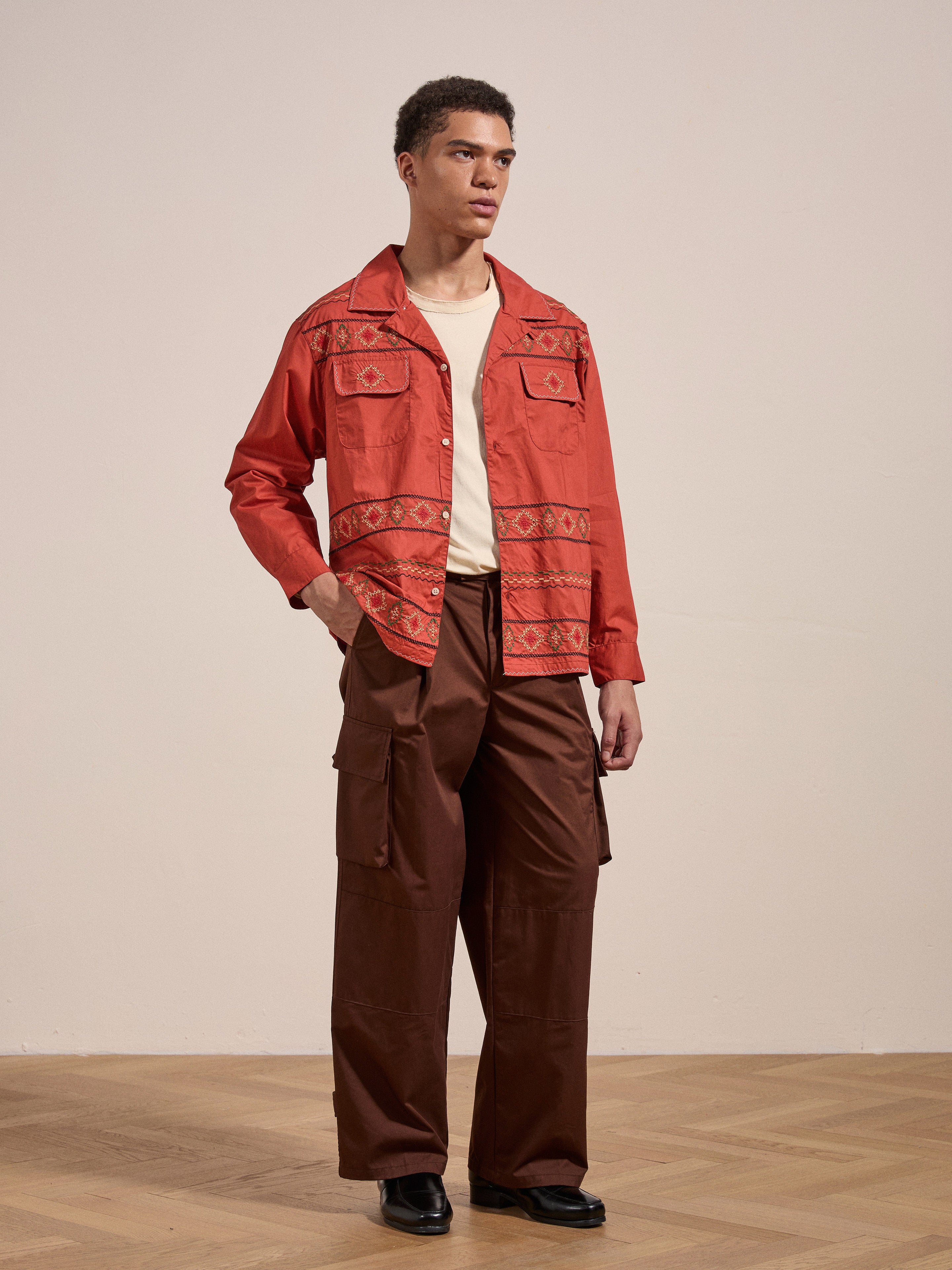 A person stands on a wooden floor in a medium-warm colored room, wearing the Embroidered Western Button Up by FOUND over a beige shirt, paired with brown cargo pants and black shoes.