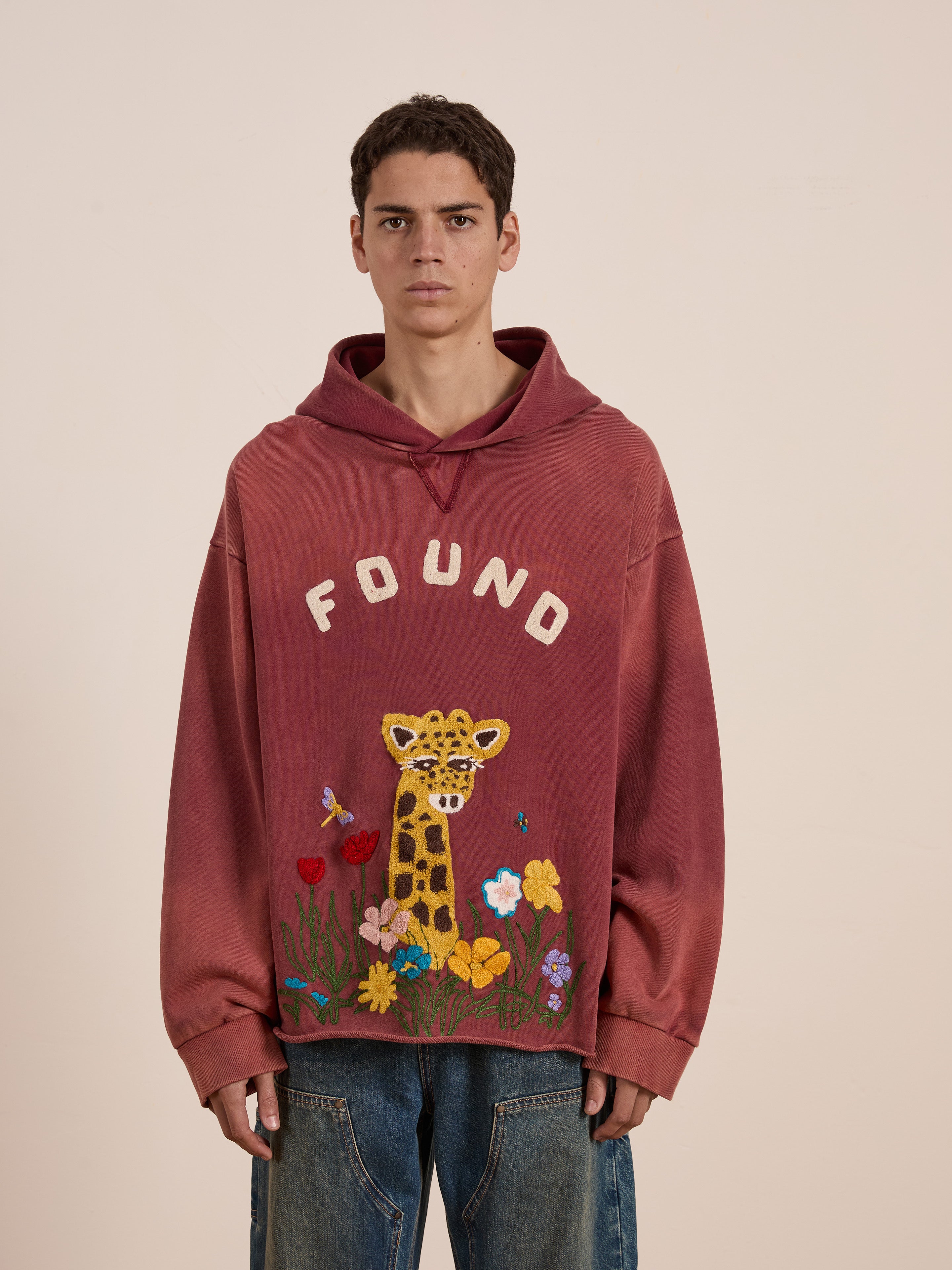 Someone is wearing the FOUND brand's Embroidered Wildlife Hoodie in red. It features a whimsical cartoon giraffe with flowers and "FOUND," combining intricate wildlife embroidery with a relaxed unisex fit for stylish comfort.