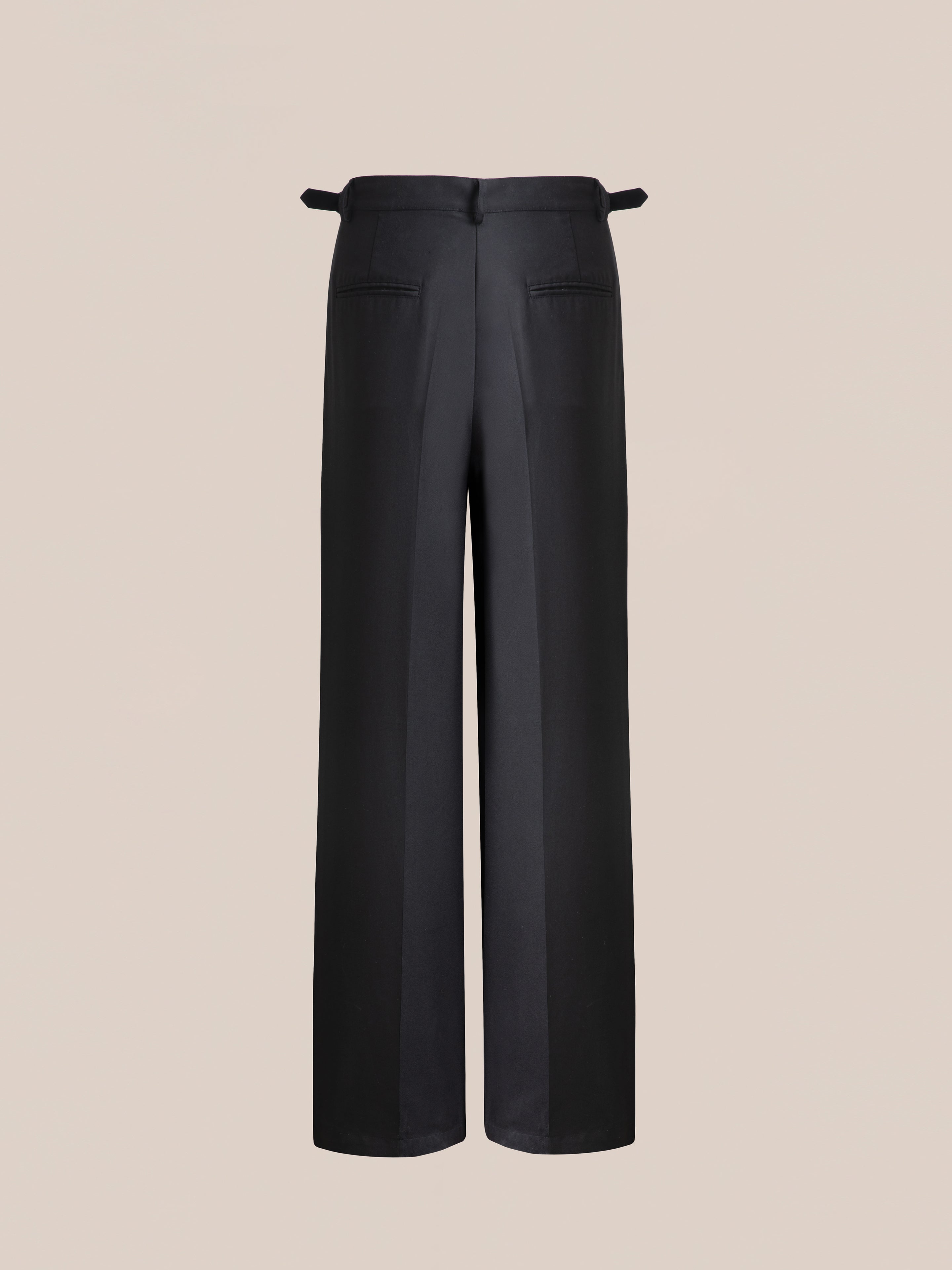 The Found Pleated Trousers, featuring a black wide-leg design with double pleats, adjustable waist tabs, and back pockets, are shown from the rear against a plain beige background.