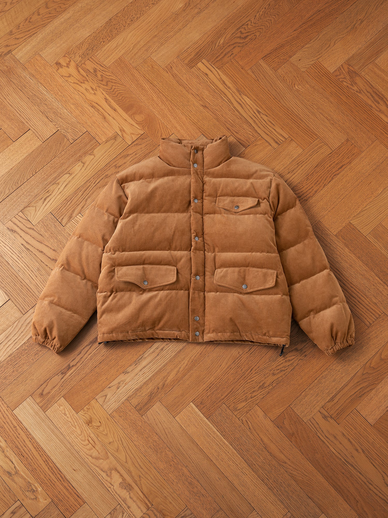 The warm tan Corduroy Puffer Jacket by FOUND features a collar, snap buttons, and front flap pockets, and is laid on a wooden herringbone-patterned floor.