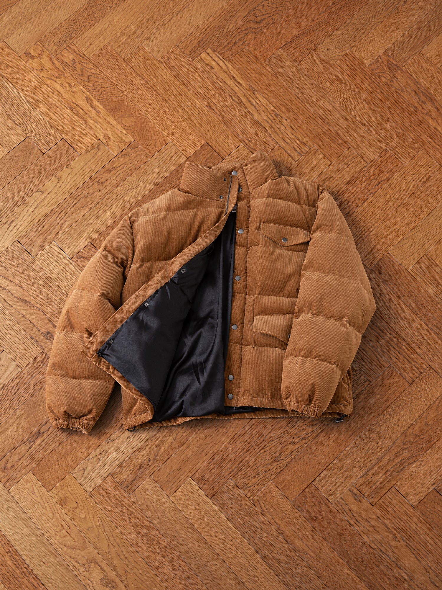 FOUND's Corduroy Puffer Jacket in warm tan with black lining and button closure, featuring front flap pockets, displayed on a wooden herringbone floor.