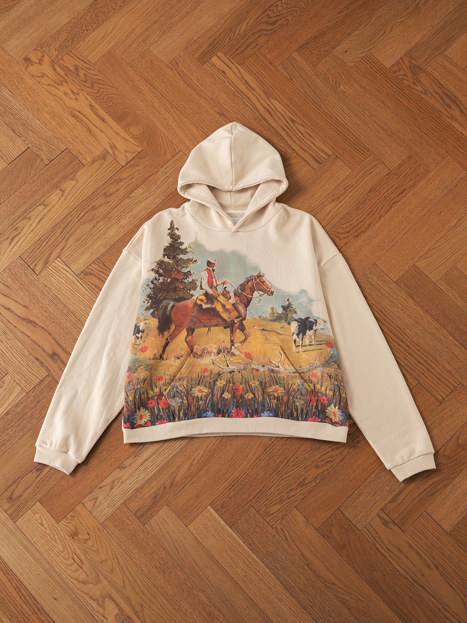 FOUND's Horseman Hoodie showcases a classic vintage style with its beige design, adorned with a cowboy on horseback among trees and cows in a grassy landscape, elegantly highlighted against a wood-patterned floor.