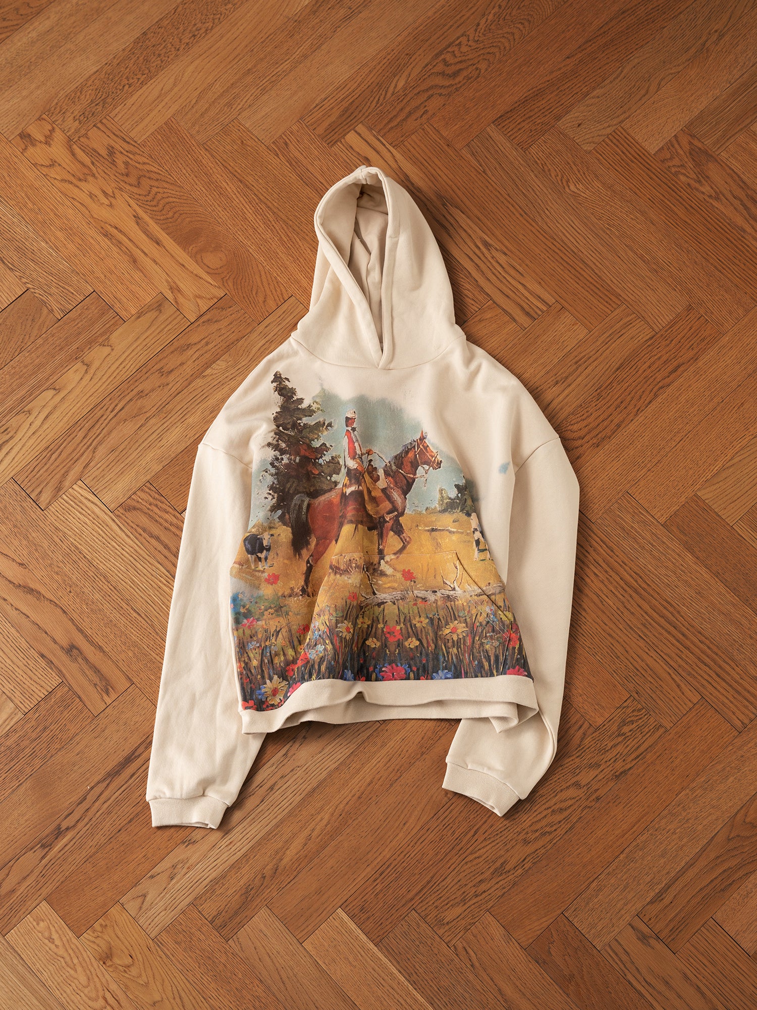 The Horseman Hoodie by FOUND, made from soft French terry cotton, showcases a detailed cowboy illustration. This beige hoodie features a depiction of a cowboy on horseback herding cattle across an open field, elegantly displayed on a wooden parquet floor.