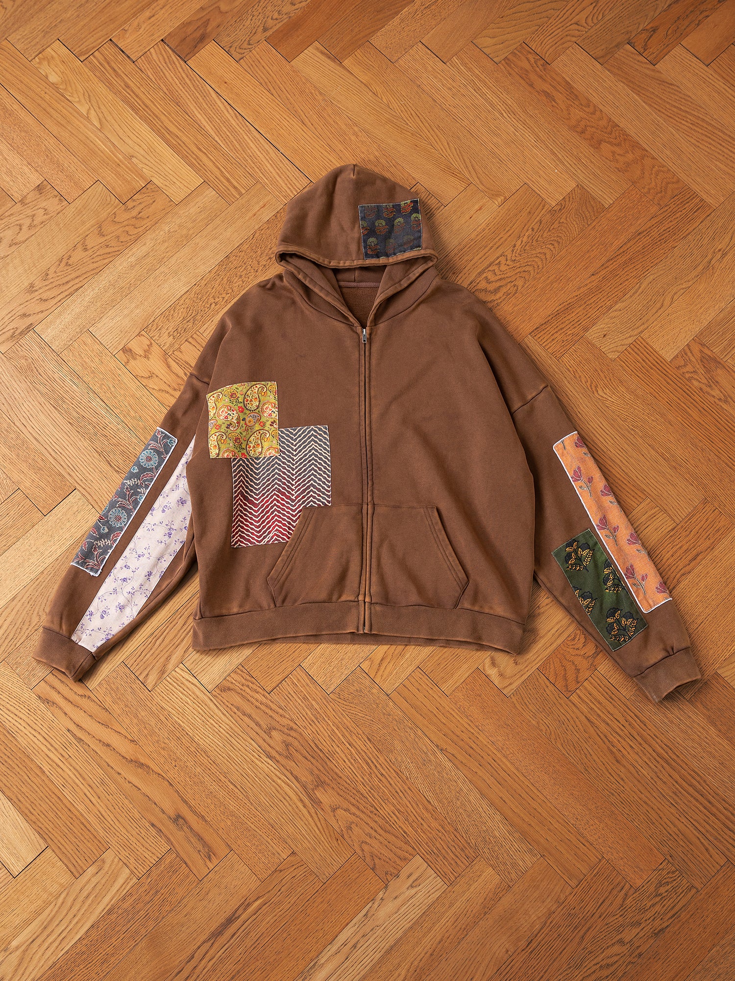 The Tapestry Patchwork Zip Up Hoodie by FOUND is displayed on a wooden floor, highlighting its vivid South Asian tapestry-inspired designs on the sleeves and chest.