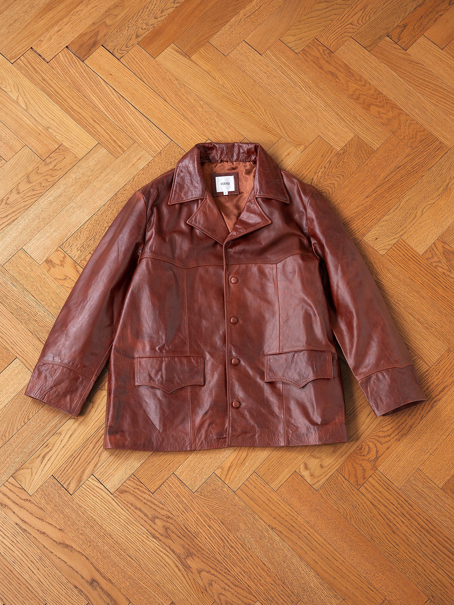 A Claret Western Leather Blazer by FOUND, made from ethically sourced lightweight cowhide, is displayed on a wooden herringbone-patterned floor, highlighting its rich brown color and large practical pockets.