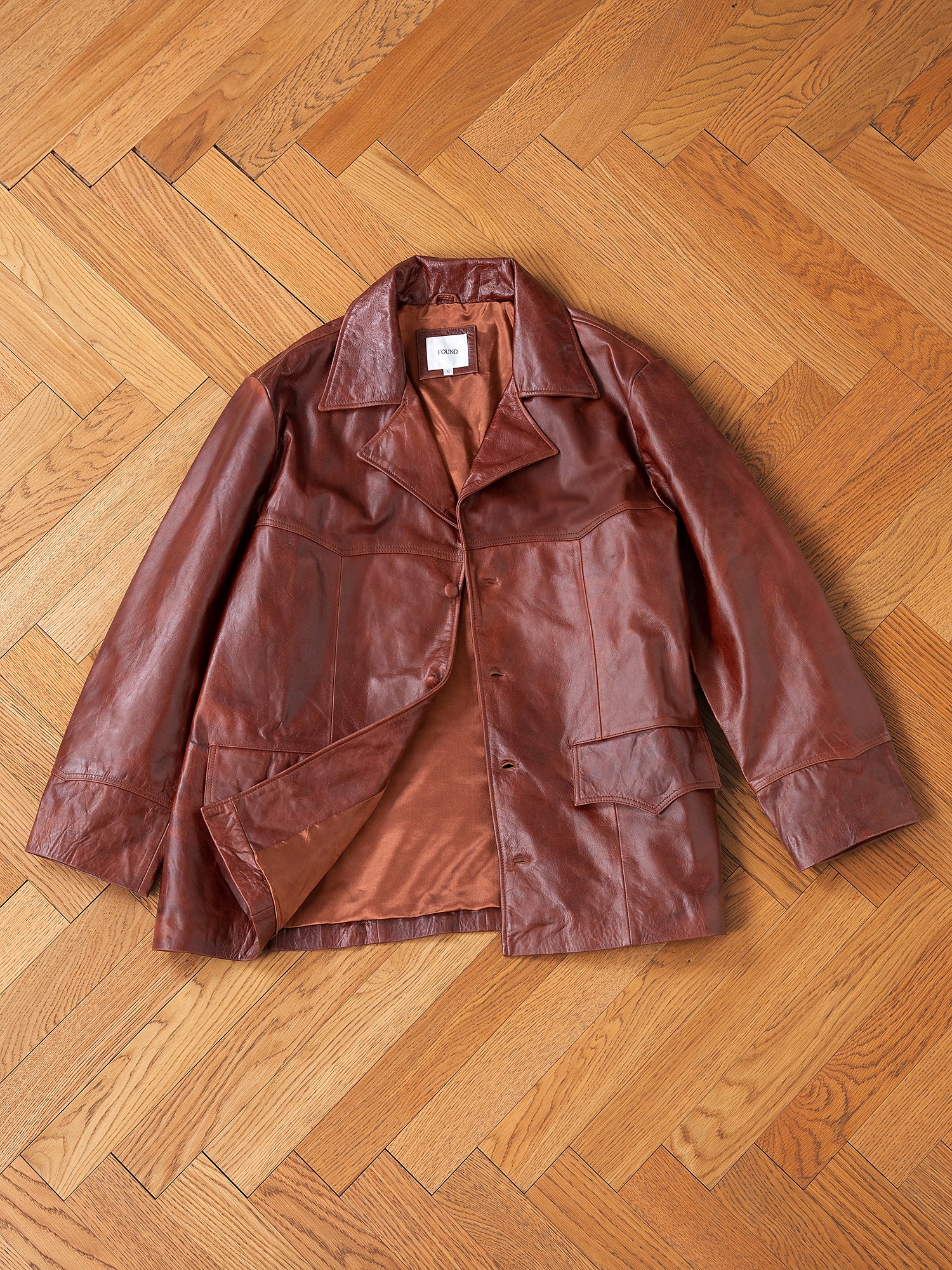 The FOUND Claret Western Leather Blazer, featuring wide lapels and front pockets, crafted from ethically made lightweight cowhide, is laid flat on a wooden herringbone-patterned floor.