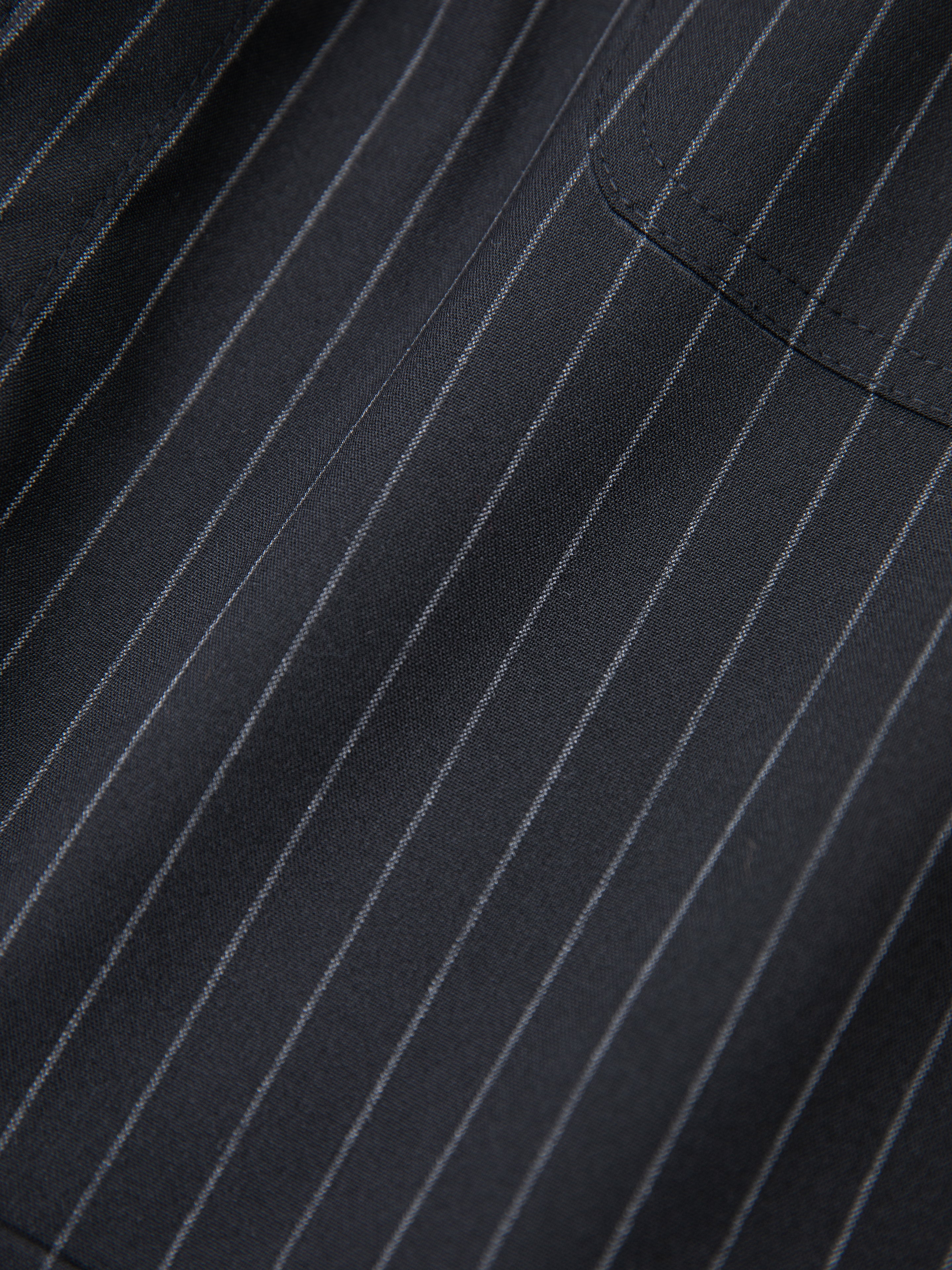 A close-up view of the Pinstripe Pleated Trousers by FOUND showcases a dark fabric with vertical thin white stripes and a partial glimpse of a pocket, elegantly suggesting their double-pleated design.