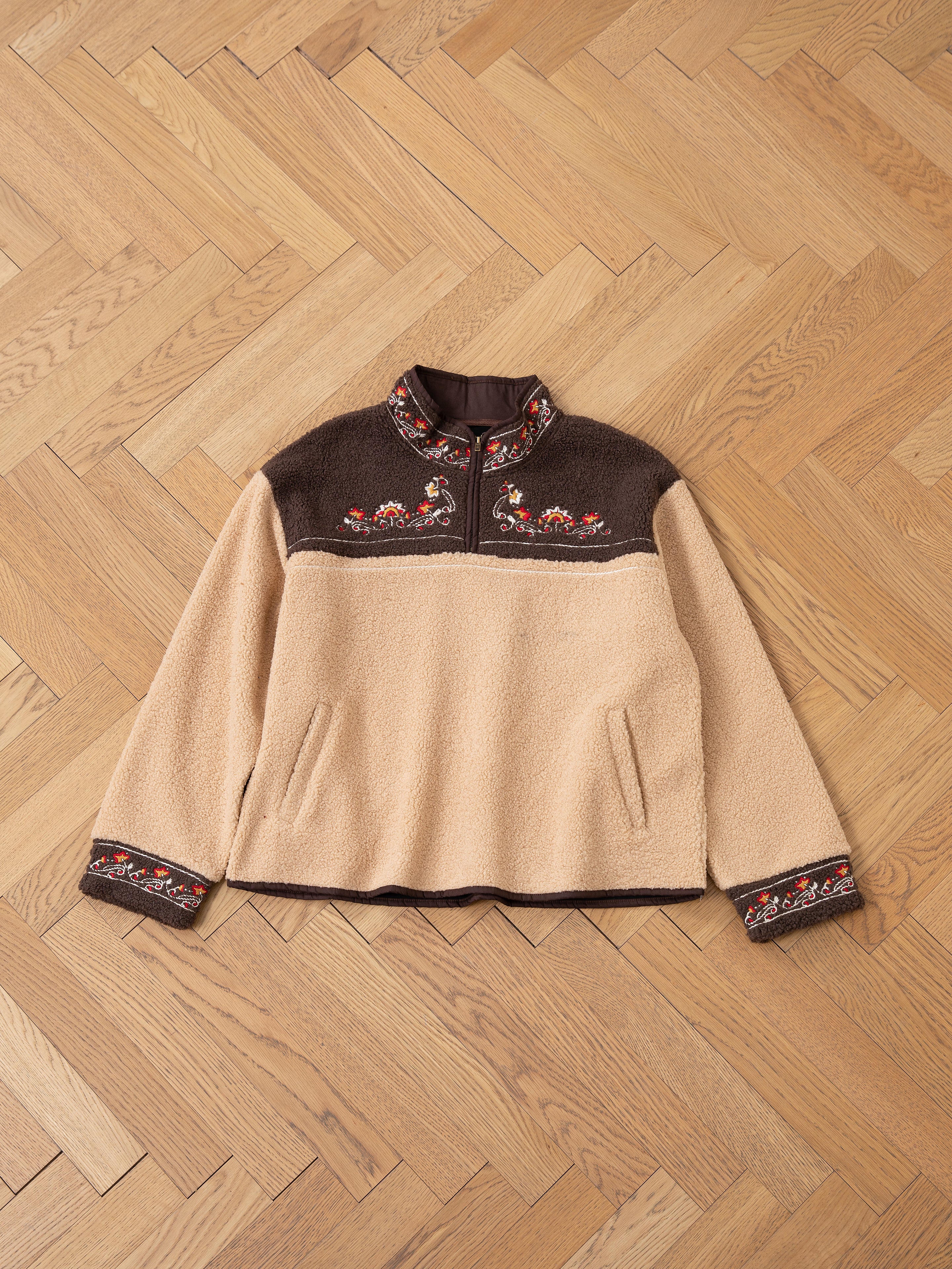 The Pahari Floral Embroidered Sherpa Pullover by FOUND, in beige and brown, showcases exquisite Pahari embroidery on the chest, cuffs, and collar, beautifully displayed on a wooden herringbone floor.