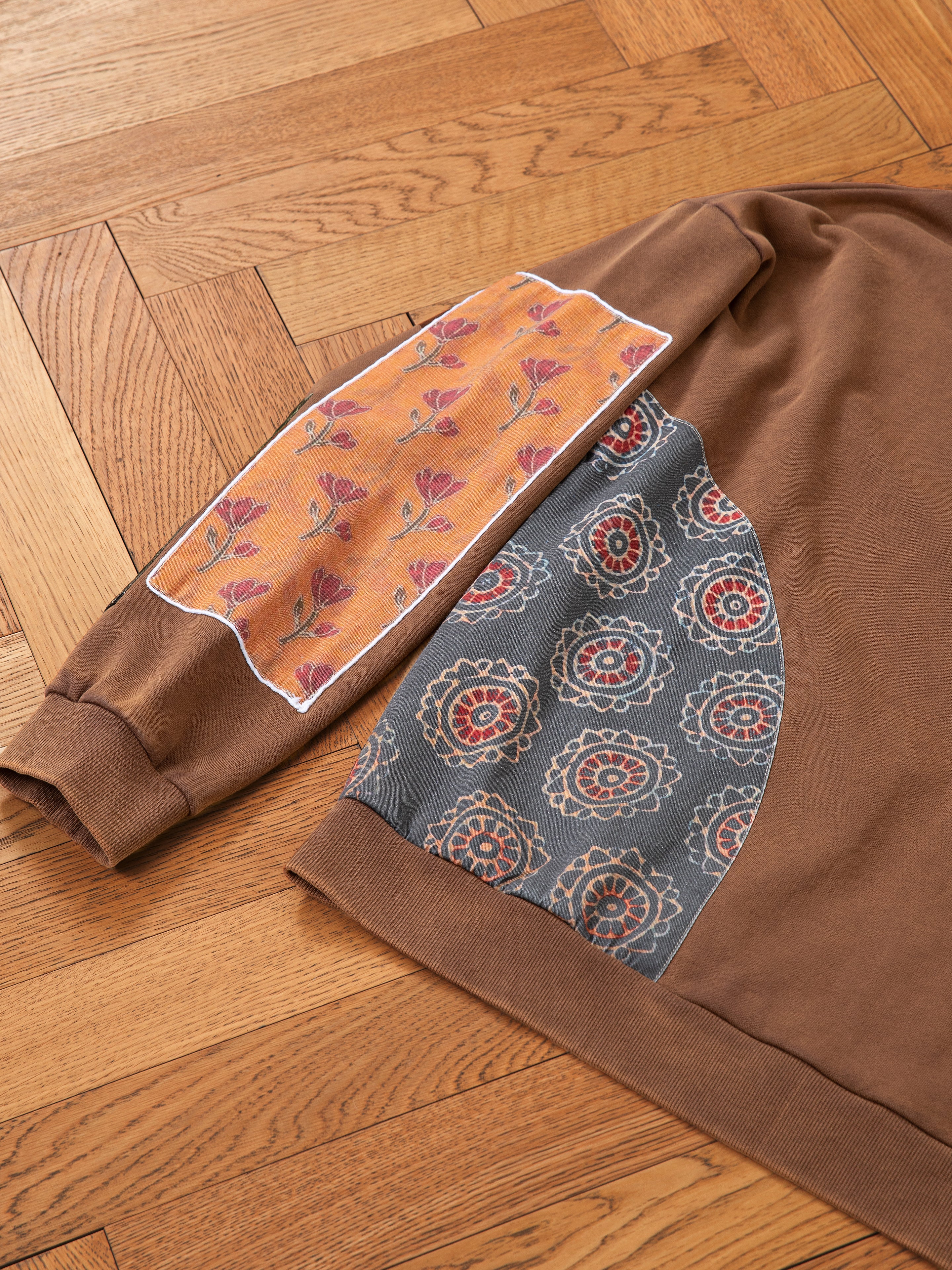 The Tapestry Patchwork Zip Up Hoodie by FOUND offers an oversized fit, featuring two distinct fabric patches: an orange floral design on the sleeve and a blue patch with circular motifs inspired by South Asian tapestry, all elegantly displayed on a wooden floor.