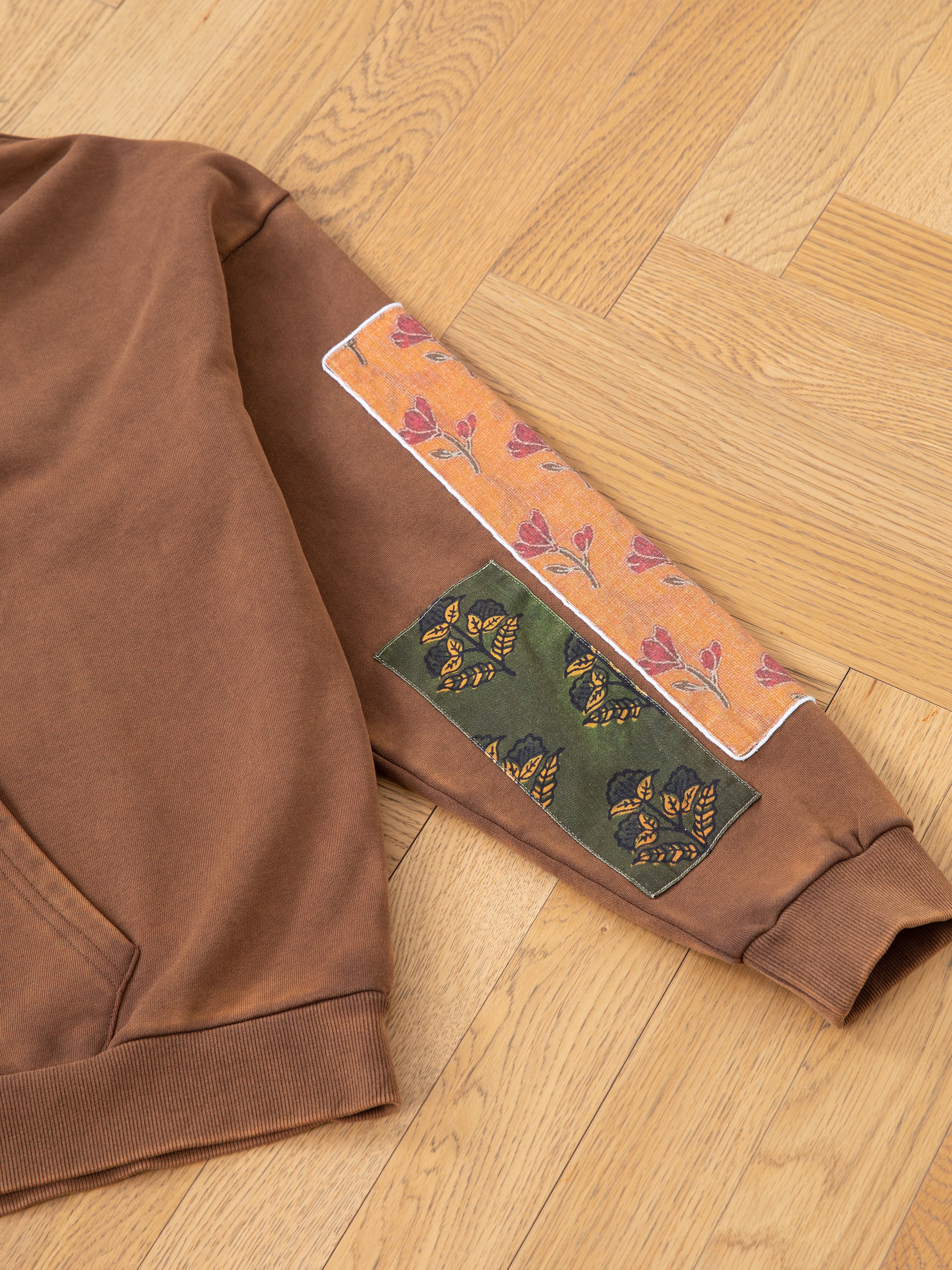 FOUND's Tapestry Patchwork Zip Up Hoodie displayed on a wooden floor, showcasing its oversized fit with detailed floral and butterfly patches on the sleeve, reminiscent of South Asian tapestry.