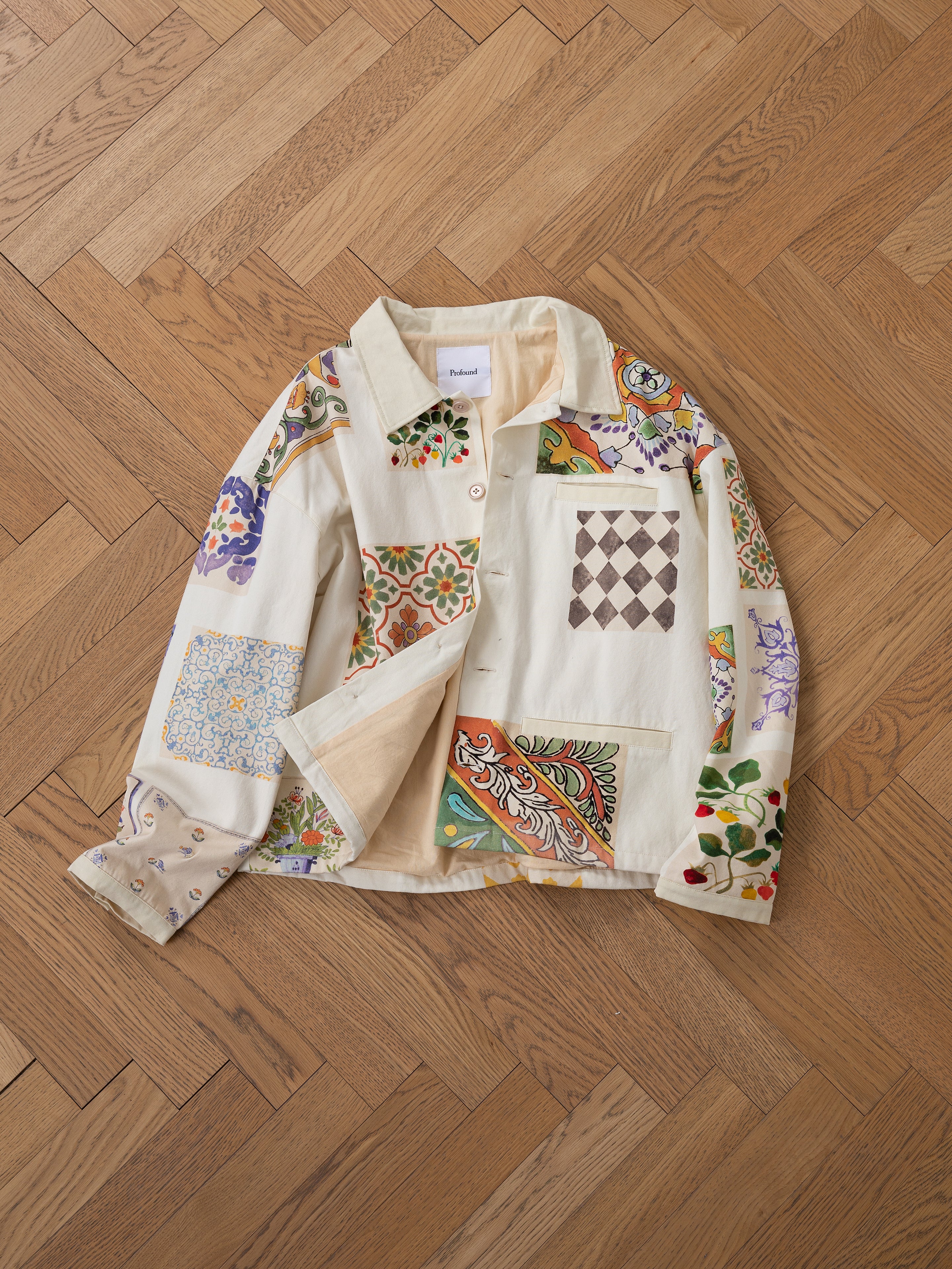 The Mosaic Work Jacket by FOUND, featuring various colorful patterns, is displayed on a wooden herringbone floor.