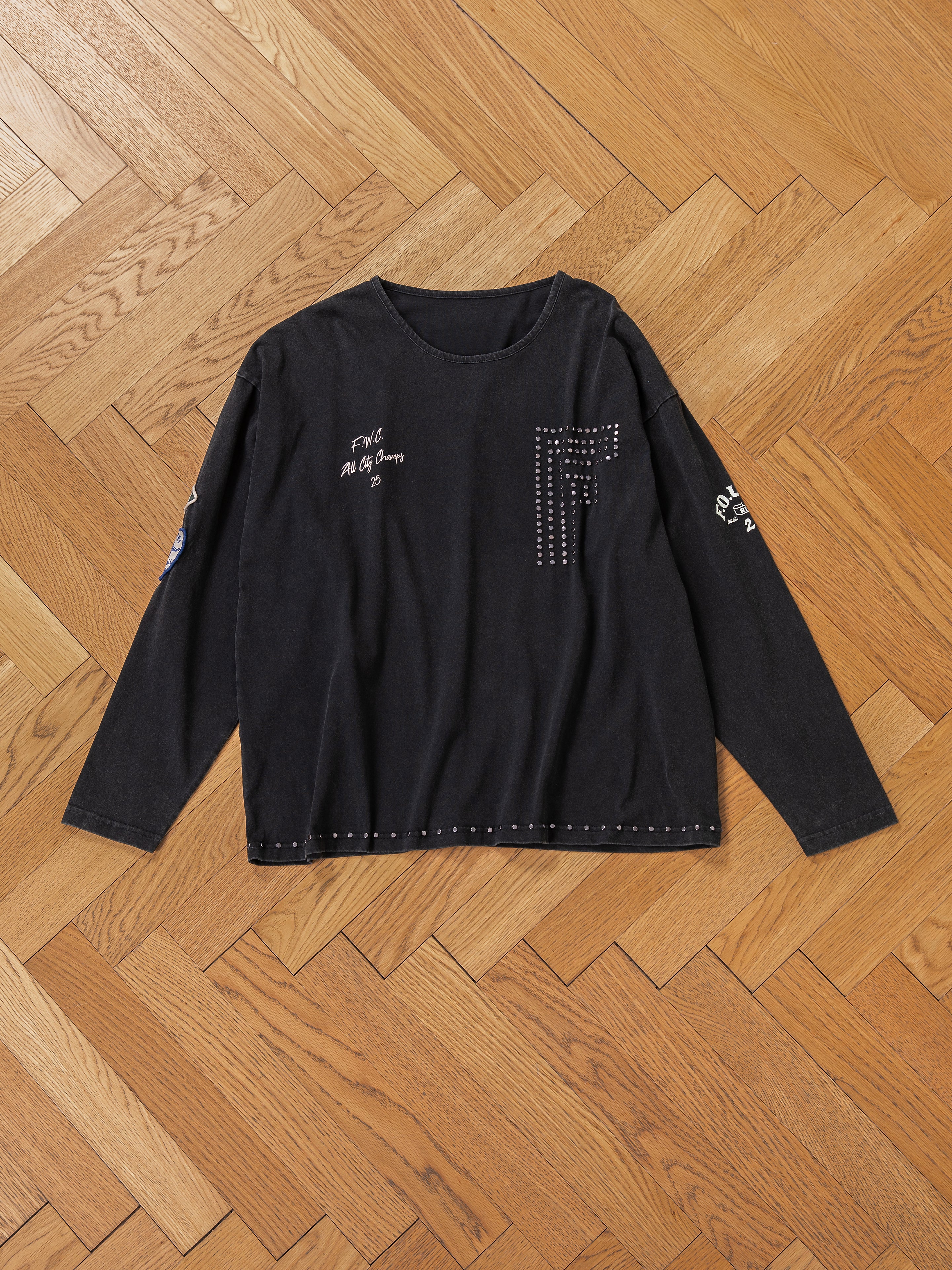 The FOUND Studded LS Shirt, a black long-sleeve adorned with decorative studs and graphic patches, is displayed flat on a parquet wooden floor.