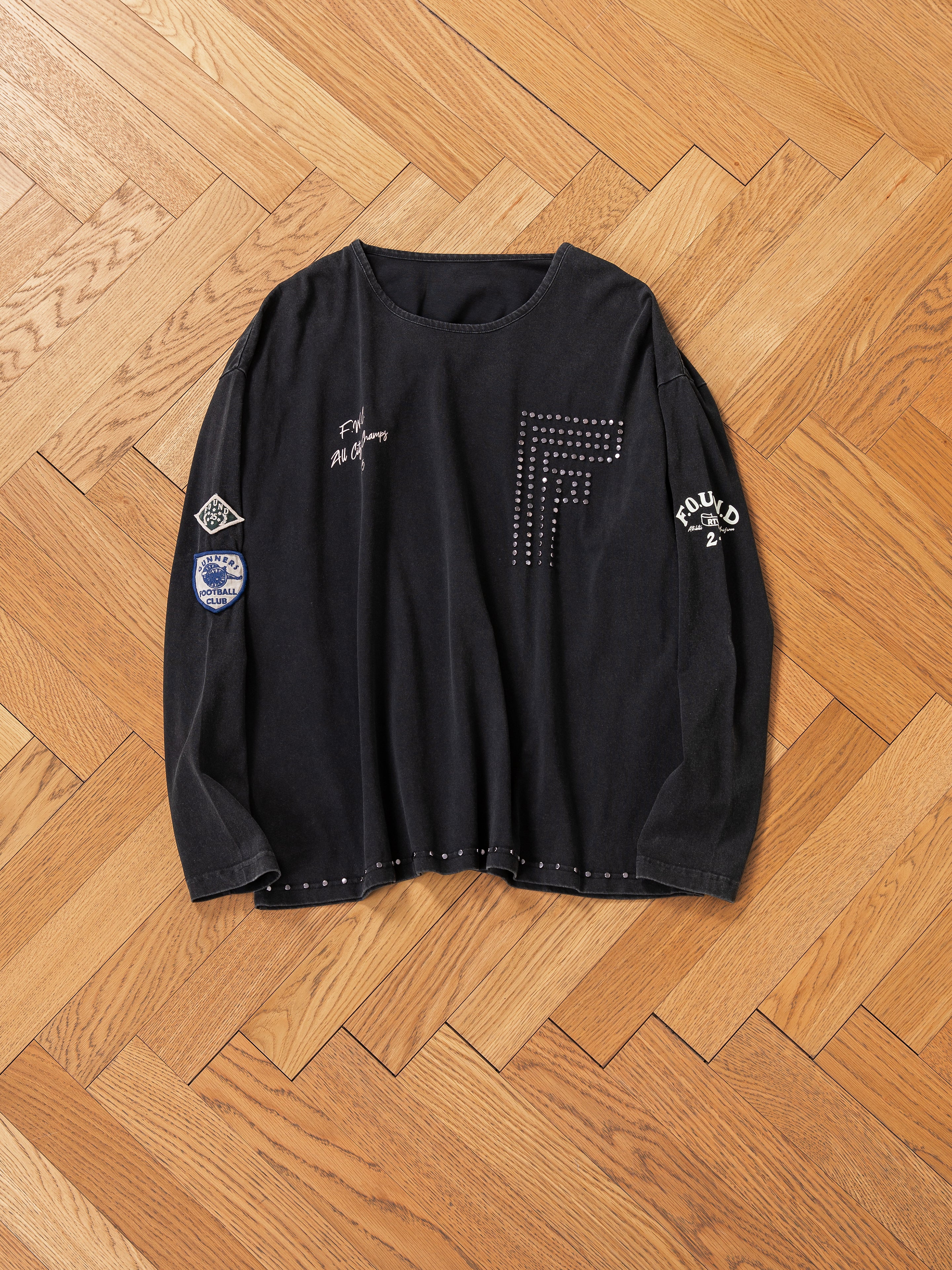 A black long-sleeve Studded LS Shirt by FOUND features various patches and a large "F" design, showcased on a wooden herringbone floor.