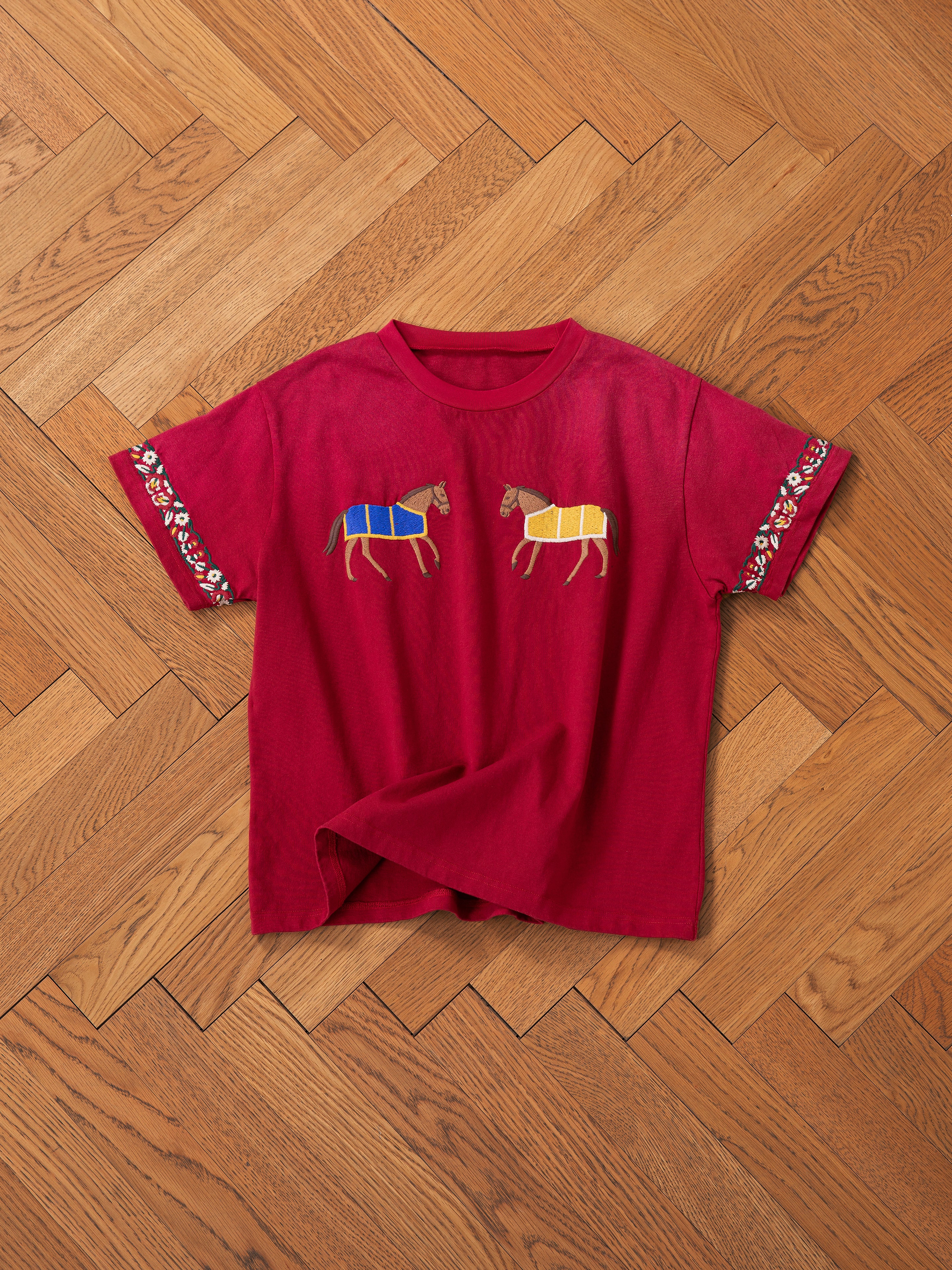 The Horse Equine Tee by FOUND is a red T-shirt, beautifully adorned with embroidered horses in vibrant racing colors on the front, highlighting blue and yellow attire. The sleeves are enhanced with floral designs. Displayed flat on a parquet floor, this T-shirt effortlessly combines style with equestrian elegance.