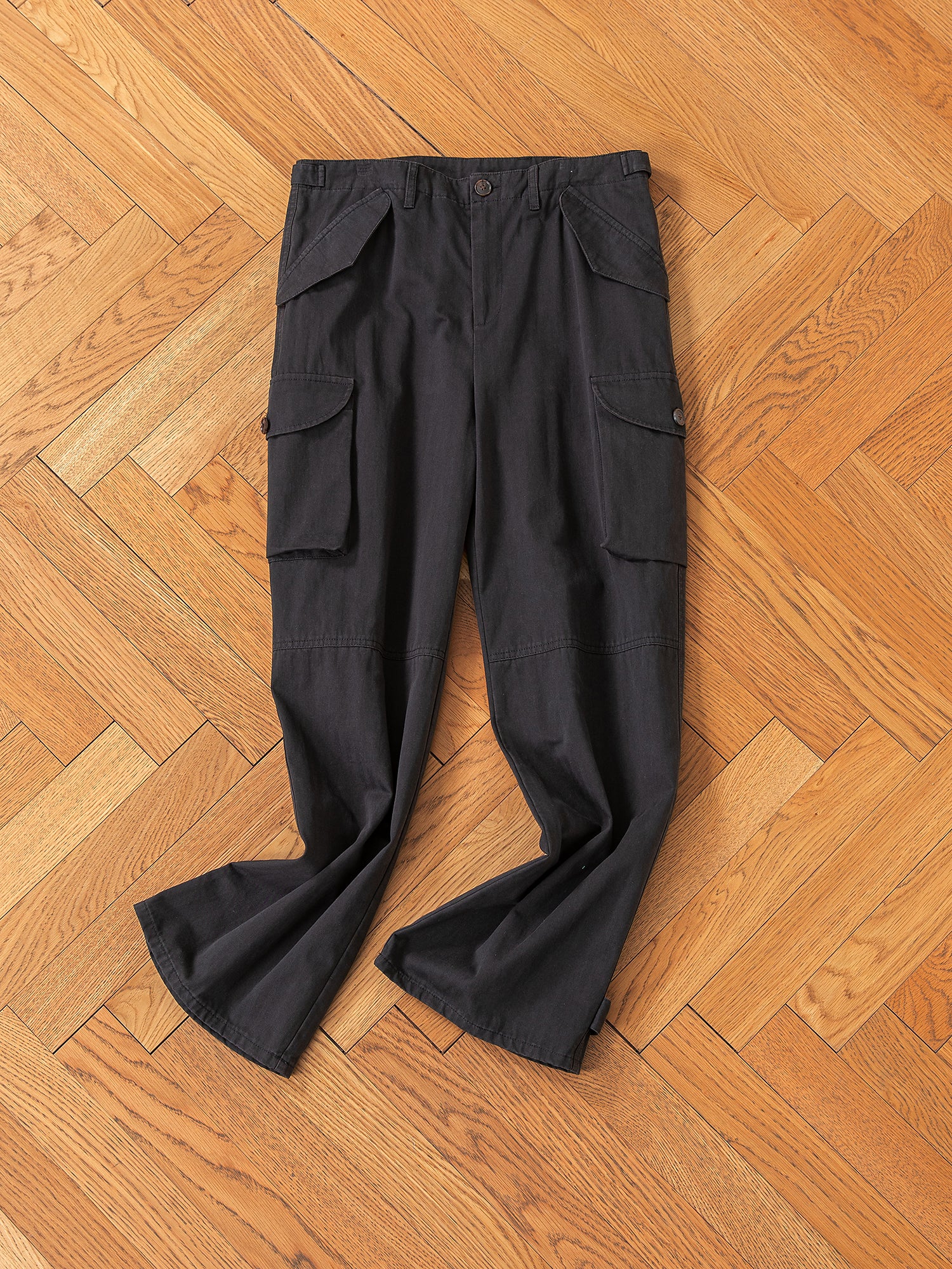 FOUND's Utility Herringbone Cargo Pants, made of durable cotton twill, feature a buttoned waist and side pockets. They offer a vintage look and robust design perfect for any adventure. Displayed on a wooden herringbone floor.