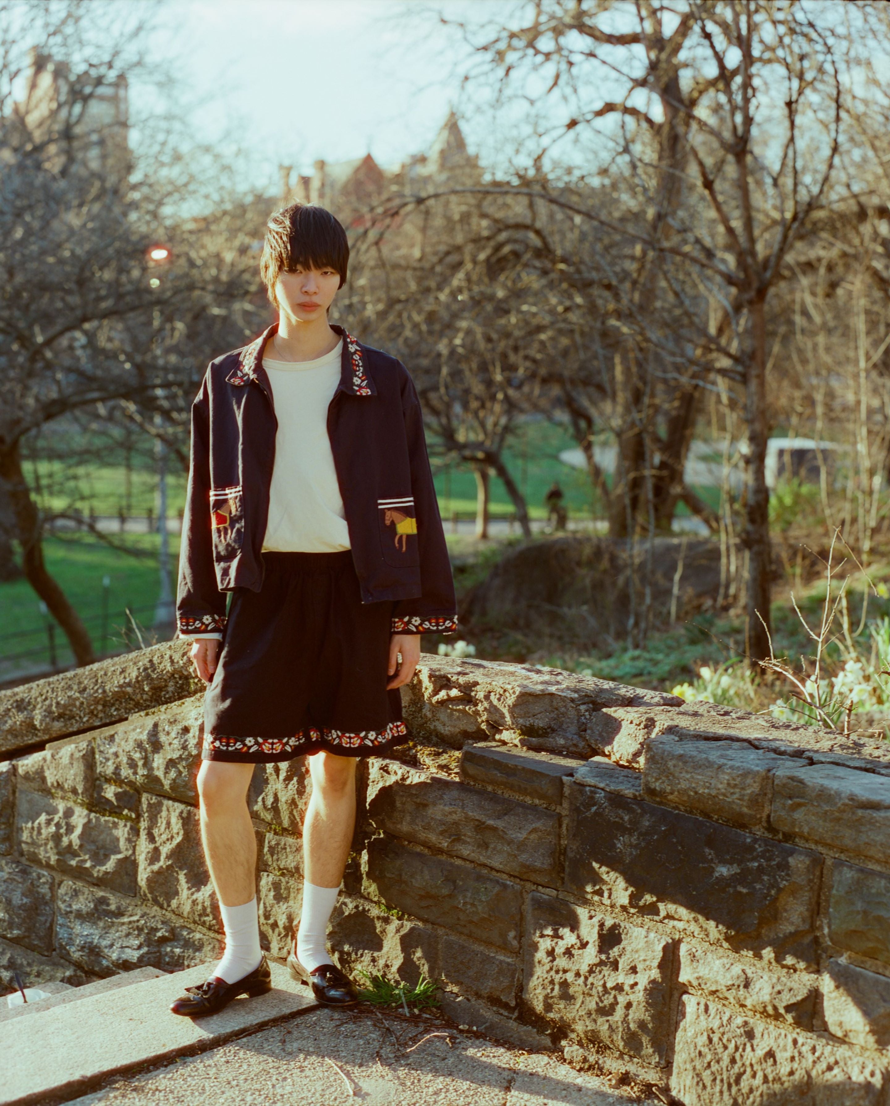 A young man stands on stone steps in a park, wearing a stylish outfit with a shirt, Horse Equine Twill Shorts by Found, and a patterned jacket, complemented by socks and loafers.