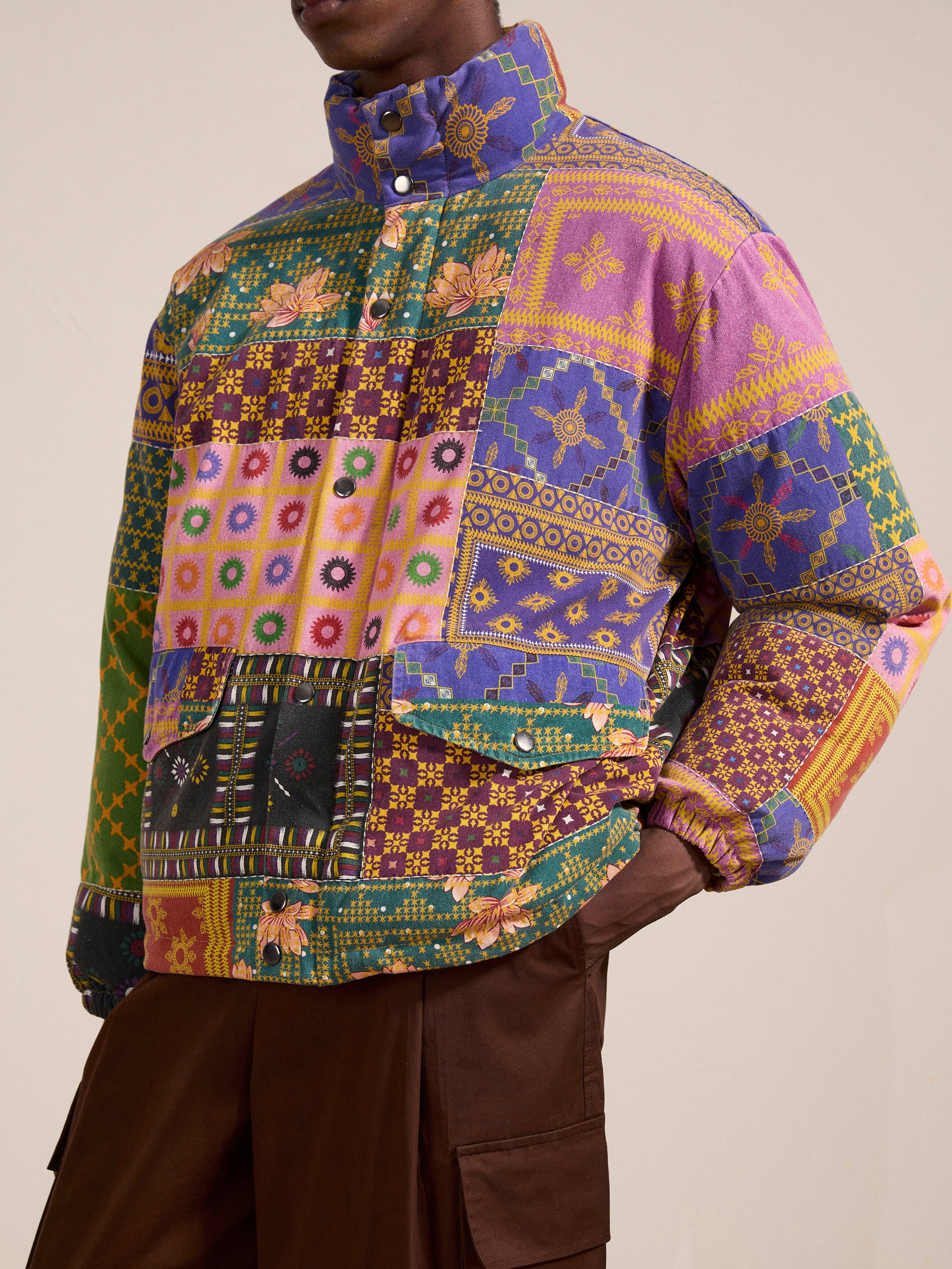 A person is wearing the Ayubia Printed Tapestry Puffer Jacket from FOUND, showcasing a high collar and detailed button accents. The jacket is adorned with intricate geometric and floral motifs in a vibrant array of colors, embodying the richness of South Asian prints. The image frames the jacket beautifully with the person's head cropped out.