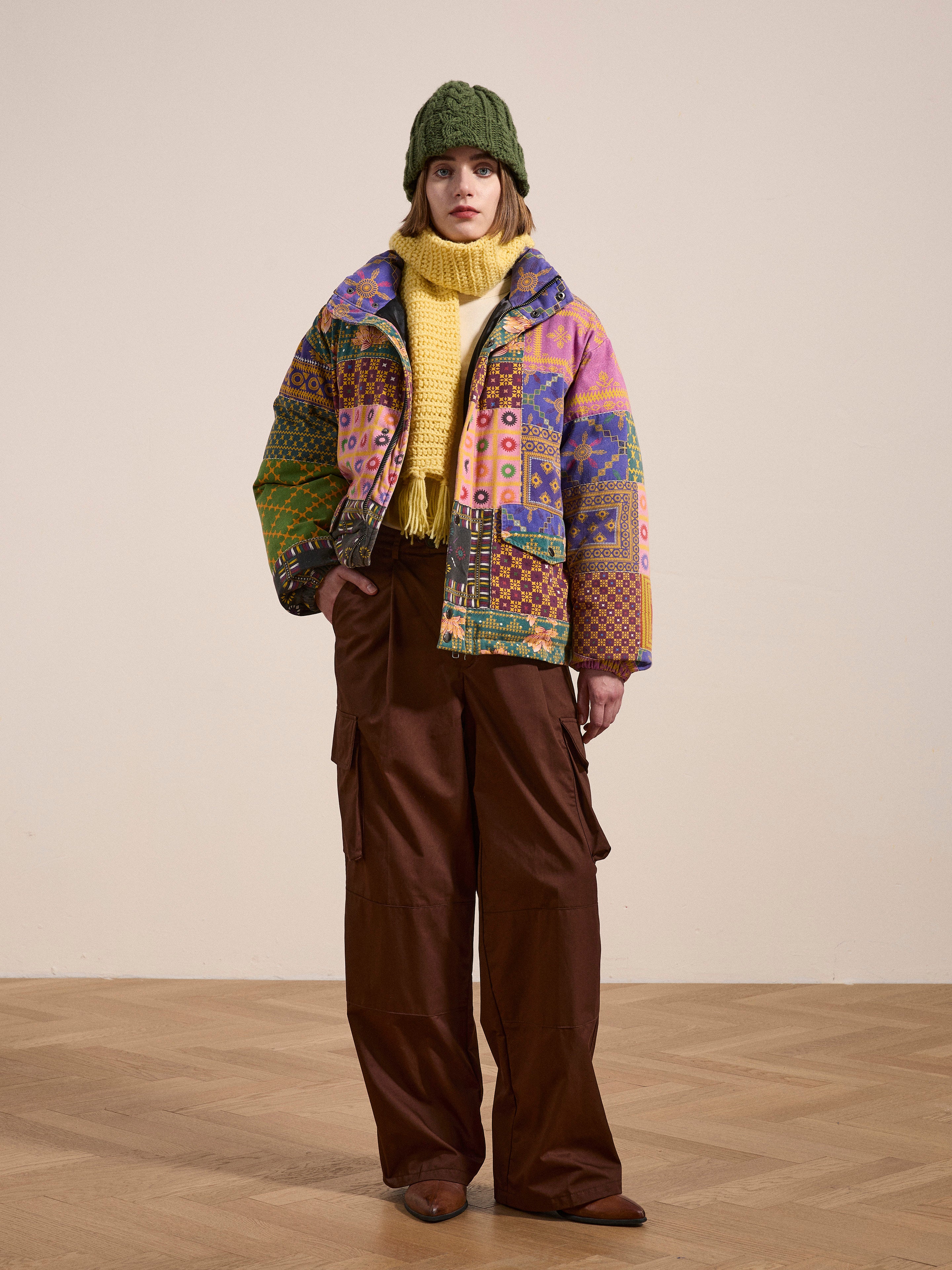 Person wearing a green knit hat, yellow scarf, and the FOUND Ayubia Printed Tapestry Puffer Jacket with vibrant South Asian prints over brown cargo pants, standing on a wooden floor against a plain beige background.