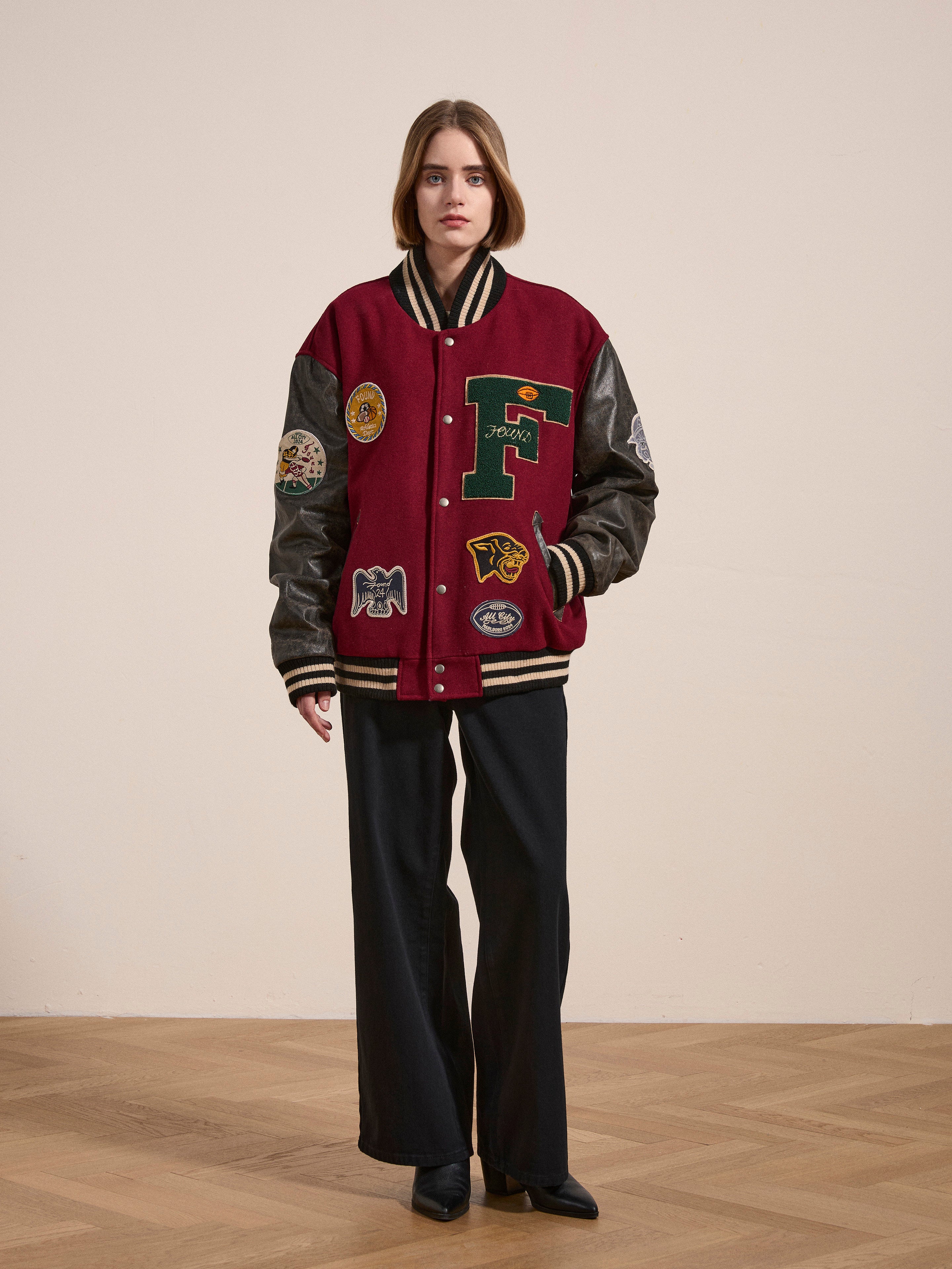 A person stands indoors wearing the Crimson Letterman Jacket from Found, adorned with various patches, over dark wide-legged pants and black shoes. The vintage look is complemented by the ethically sourced materials used in the design. The background is plain and light-colored.