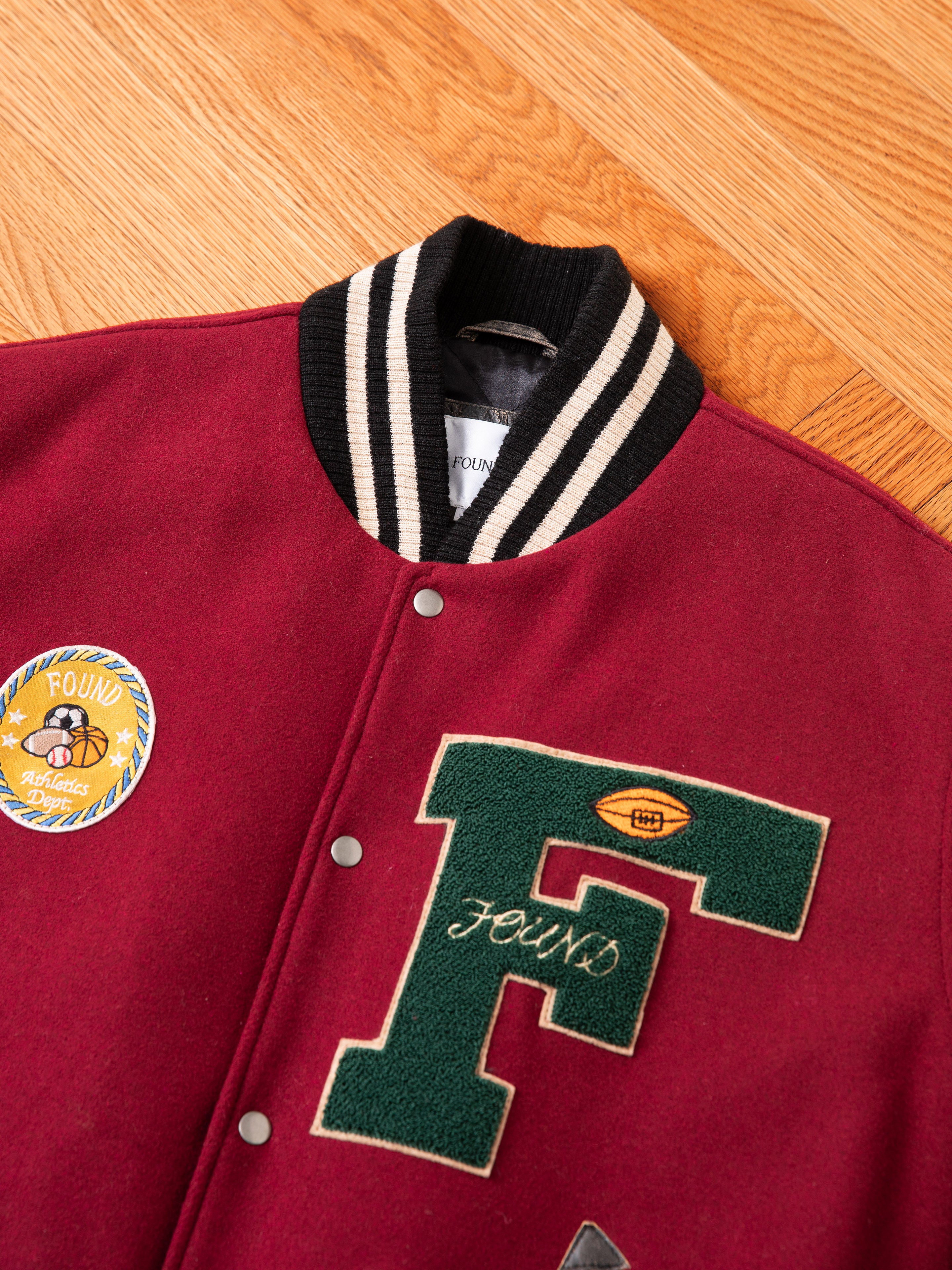 A Crimson Letterman Jacket by Found, made from ethically sourced materials, showcases a maroon color with an "F" patch on the front and a circular patch on the chest, displayed on a wooden floor for that perfect vintage look.