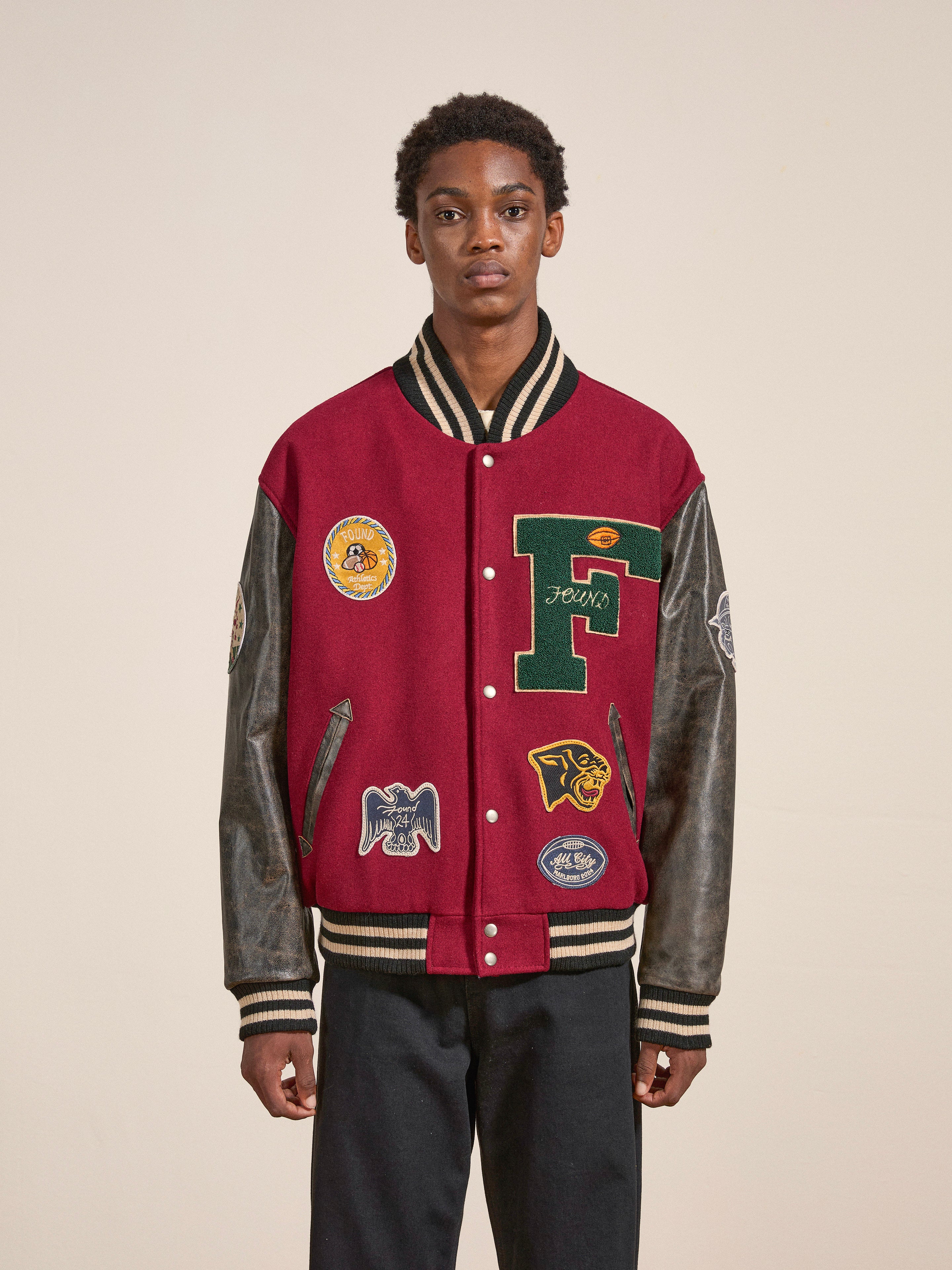 A young individual stands facing forward, donning a red and black Crimson Letterman Jacket from Found, adorned with multiple patches, paired with dark pants against a plain backdrop. The jacket, made from ethically sourced materials, boasts a vintage look that is both timeless and stylish.