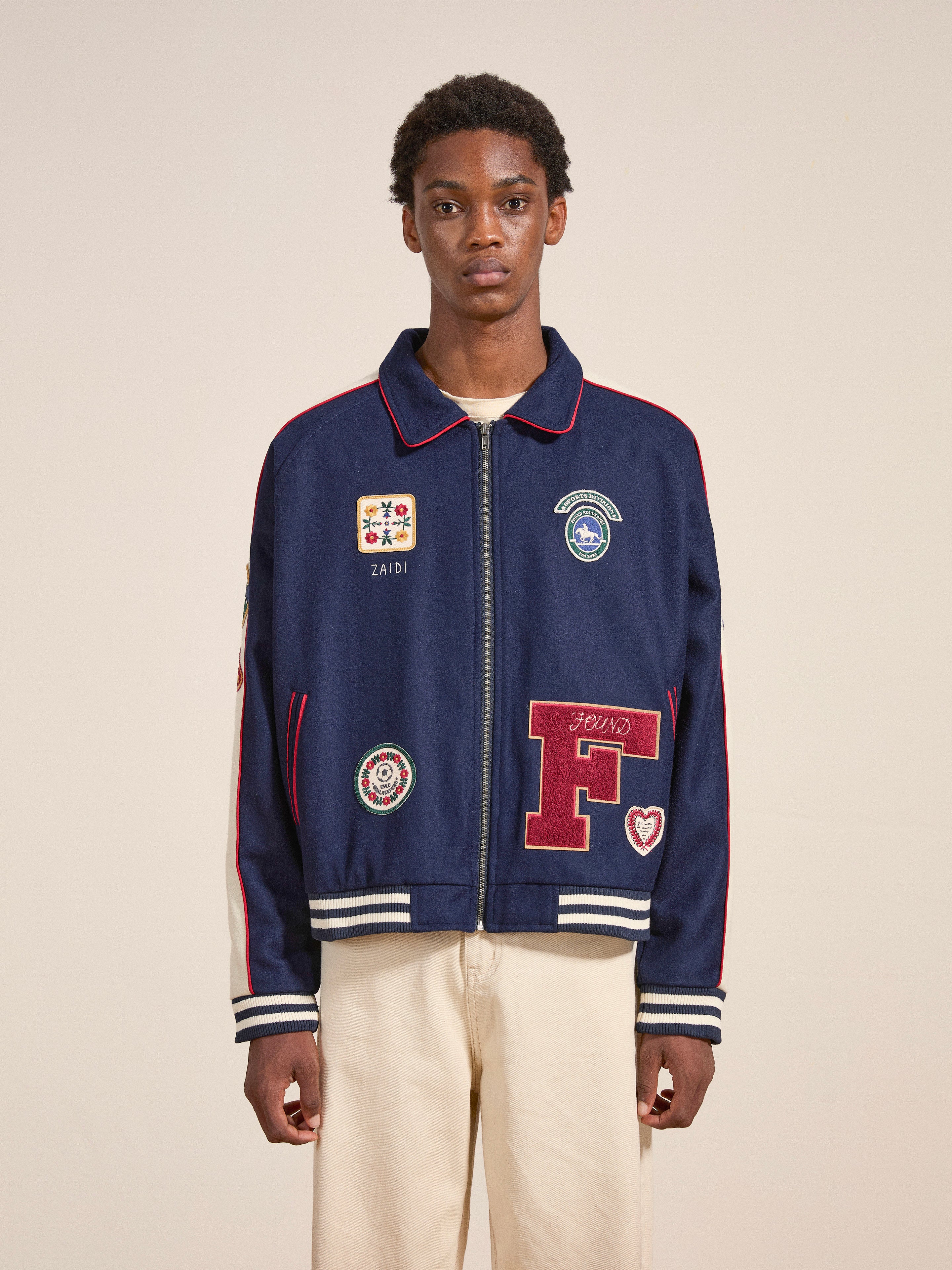 A person stands against a plain background, showcasing the mid-weight wool blend FOUND All Roads Varsity Jacket adorned with various patches that exude a vintage look, paired with light-colored pants.