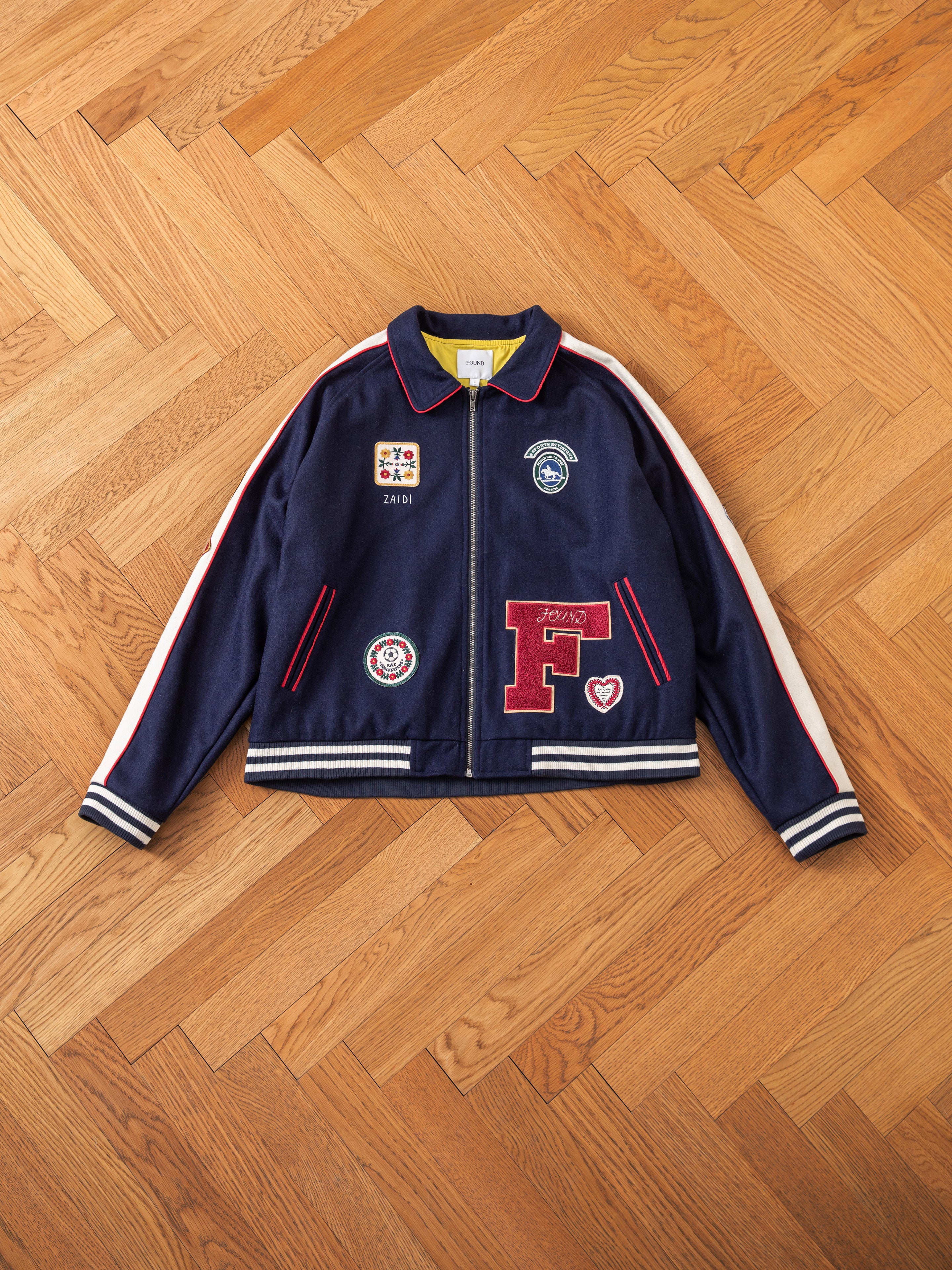 The All Roads Varsity Jacket by FOUND is crafted from a mid-weight wool blend and showcases a blue fabric accented with red and white stripes on the sleeves. The jacket also features multiple patches on the front and a central zipper, exuding a vintage look as it rests on a herringbone wooden floor.