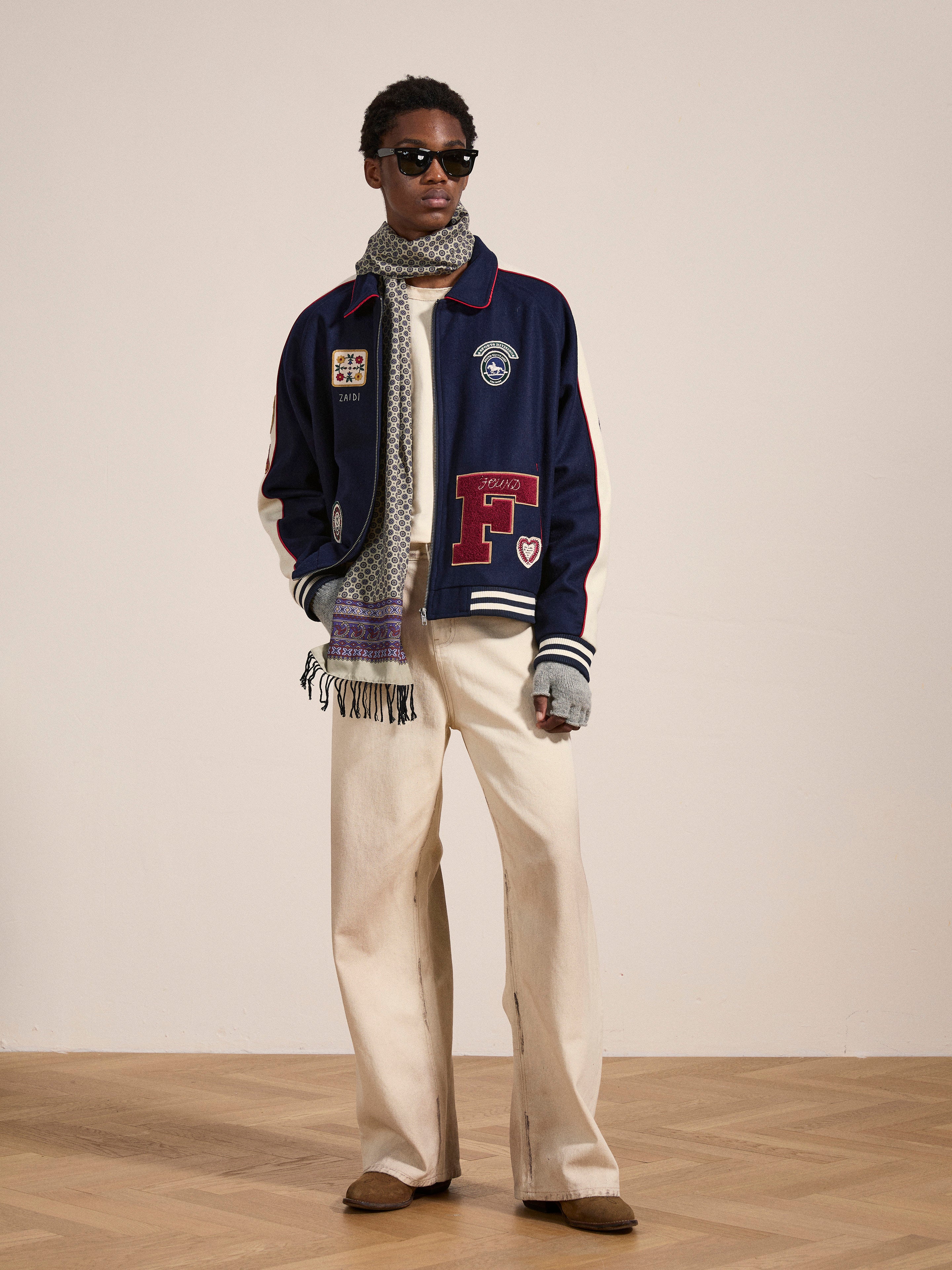 A person stands against a plain background, donning sunglasses, a blue All Roads Varsity Jacket with patches by FOUND, a patterned scarf, white wide-leg pants, and brown shoes. The mid-weight wool blend jacket enhances the vintage appeal of the outfit.