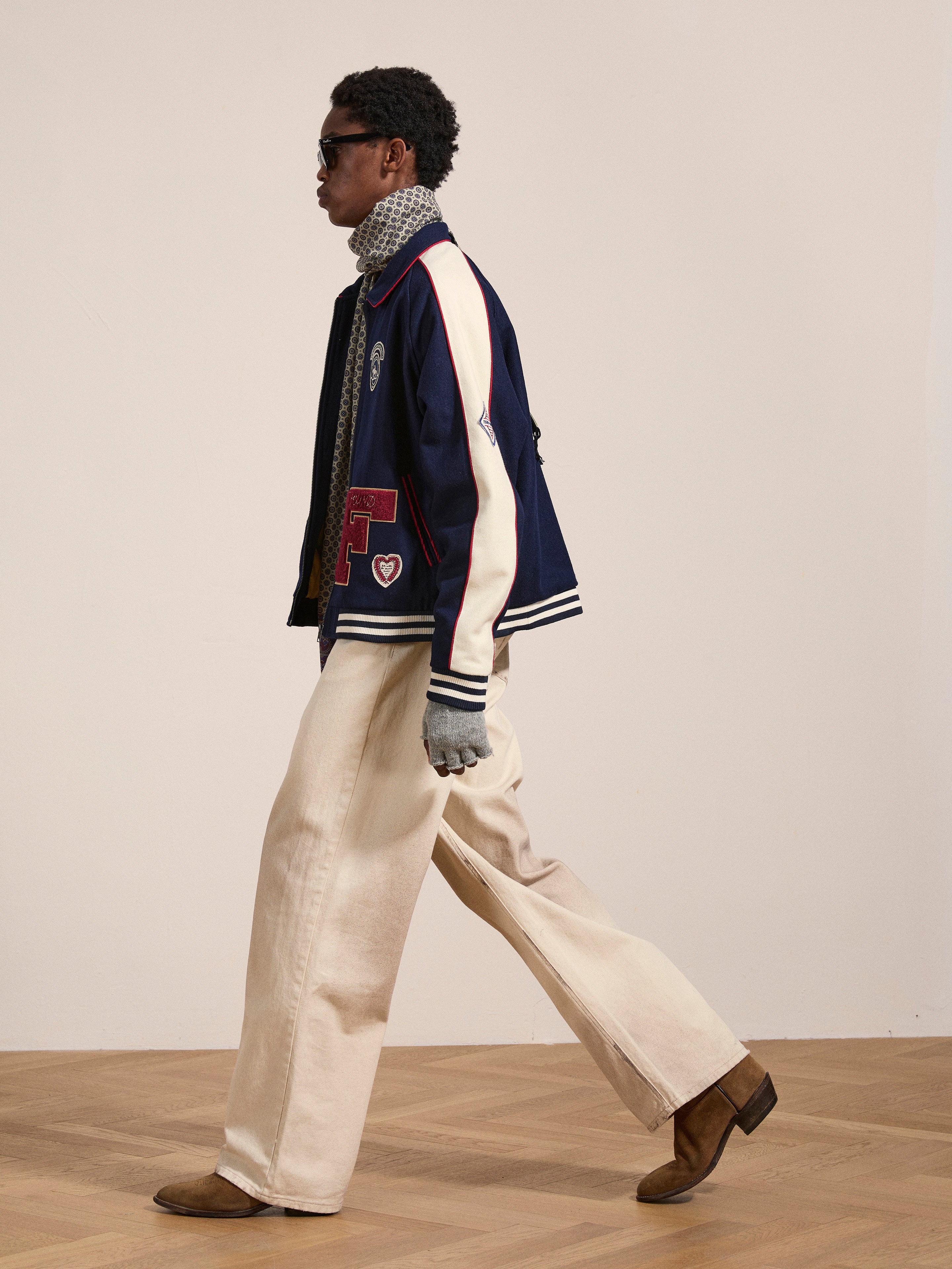 A person in a FOUND All Roads Varsity Jacket with white sleeves, a gray scarf, beige wide-legged pants, brown boots, and dark sunglasses walks across a wooden floor. The dark blue mid-weight wool blend jacket gives a vintage look against the plain light wall backdrop.