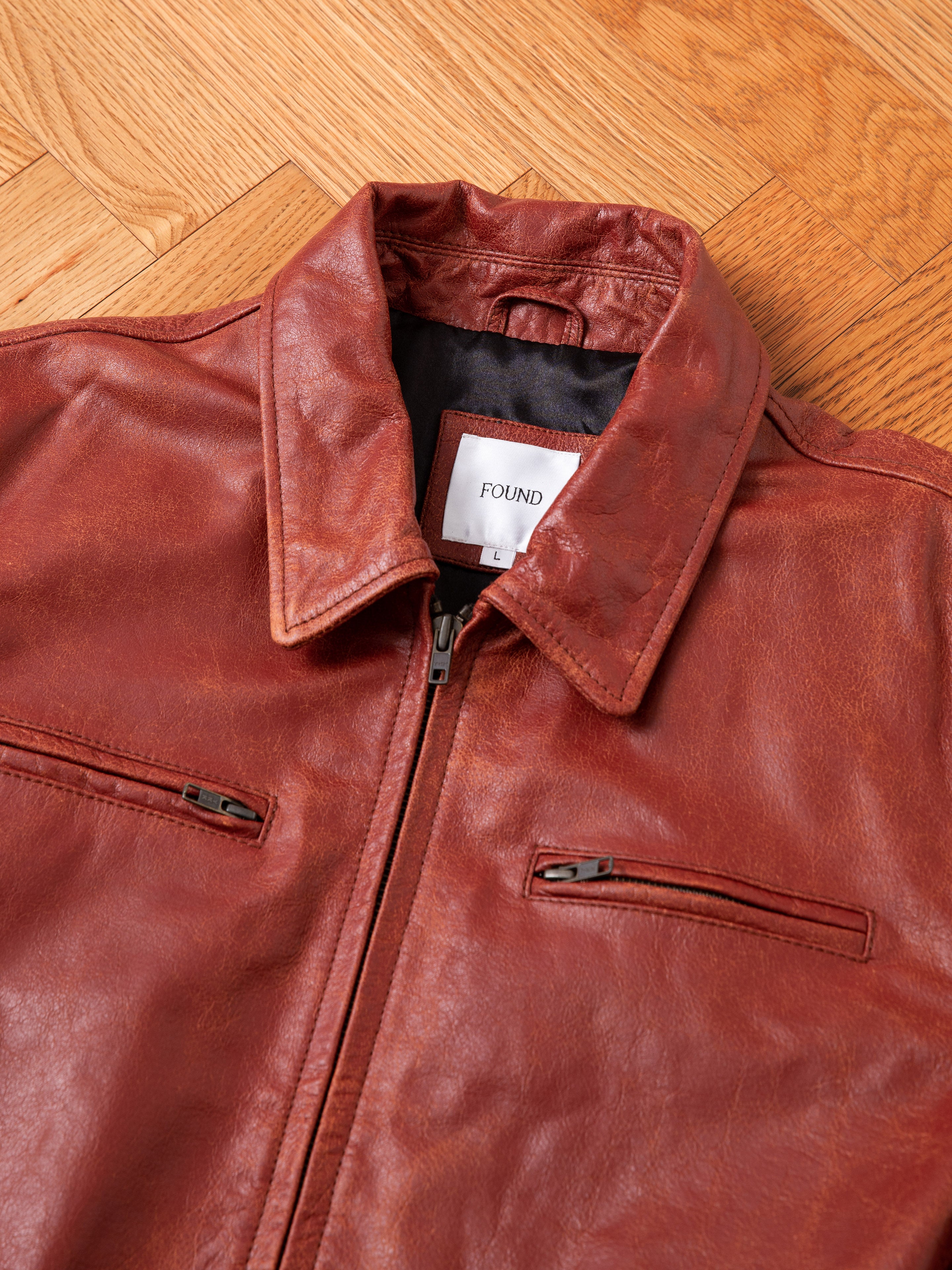 A brown distressed Marlboro Sun Faded Leather Racer Jacket with a zip-up front and two zippered chest pockets is laid flat on a wooden herringbone floor. A label inside the jacket reads "FOUND.