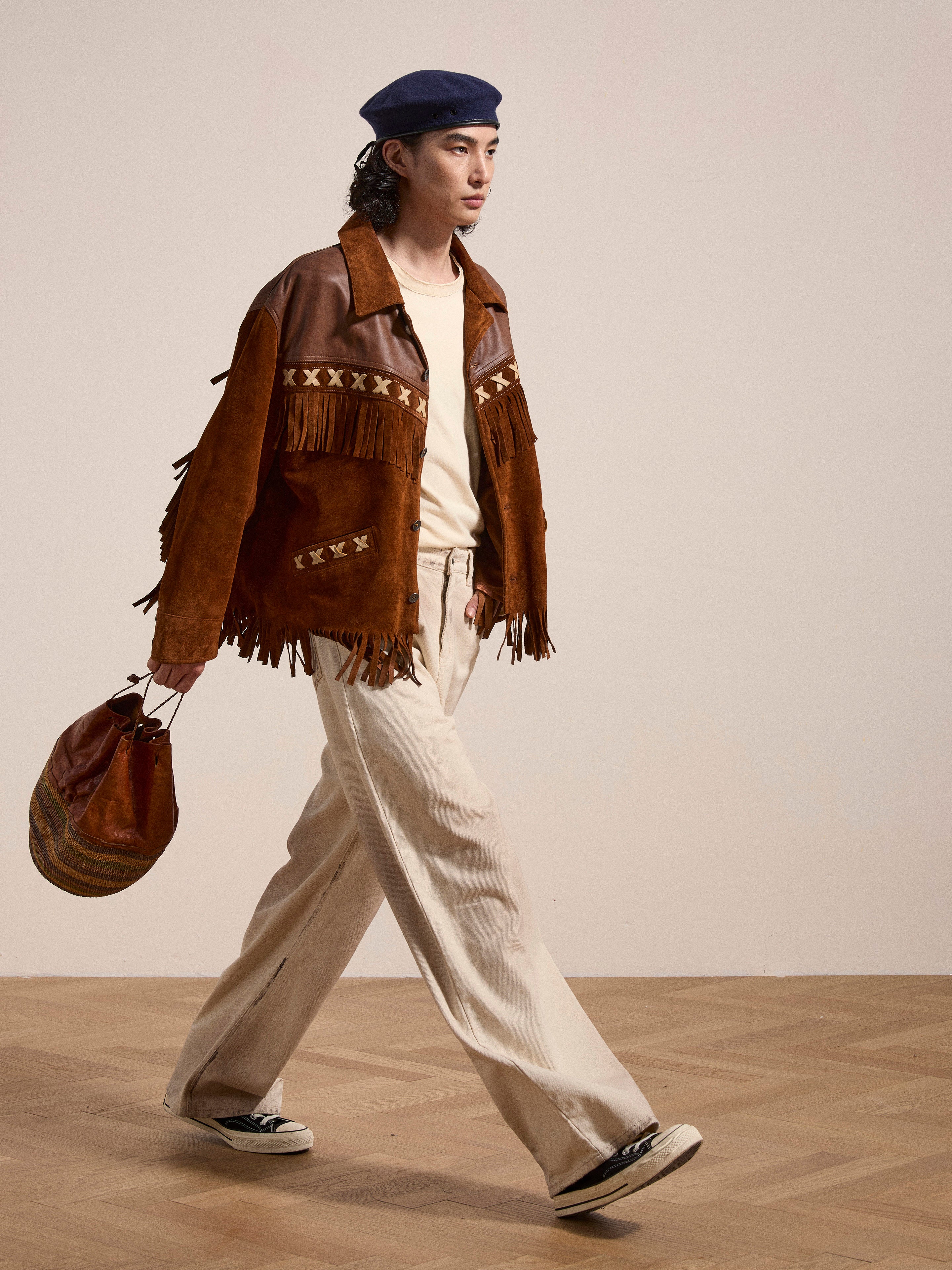 A person confidently strides wearing FOUND's Western Fringe Suede Leather Jacket, paired with cream pants and a navy beret, holding a stylish brown bag.