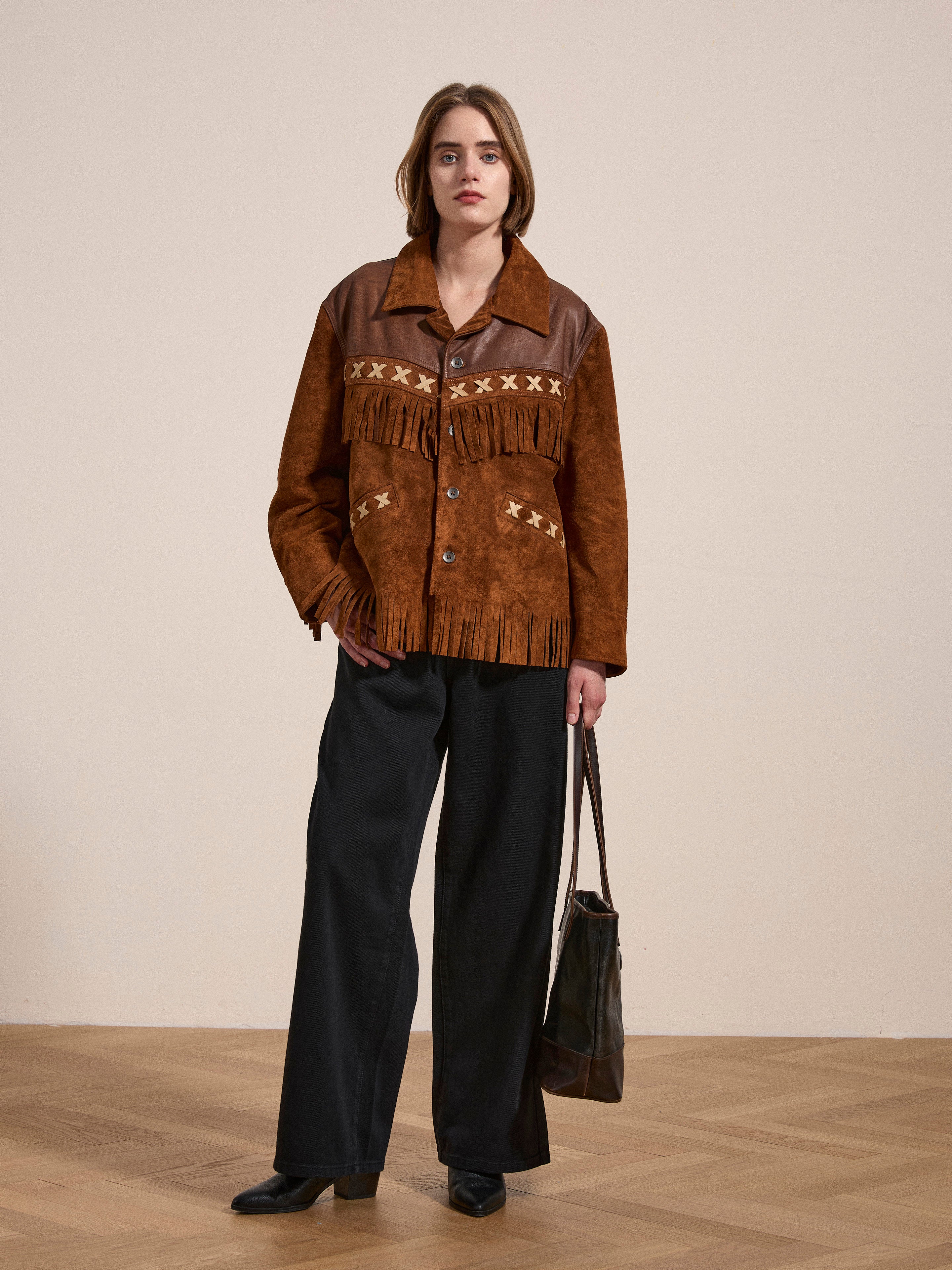 A person wearing a FOUND Western Fringe Suede Leather Jacket with patterned details, paired with black pants and black shoes, holds a brown ethically sourced bag while standing on a wooden floor.