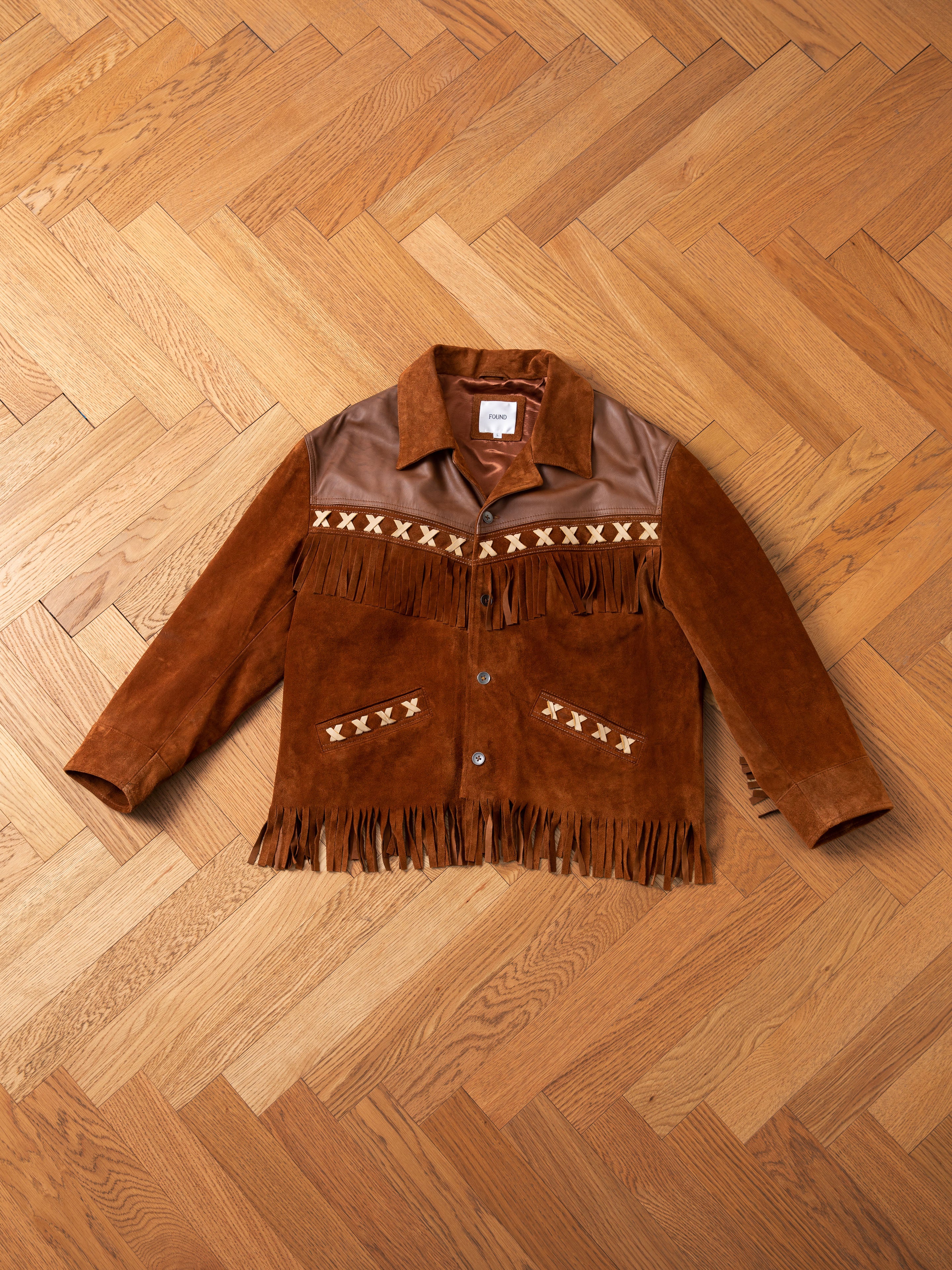 The FOUND Western Fringe Suede Leather Jacket is made from genuine suede, featuring decorative X patterns on the chest and pockets, shown laid flat on a wooden floor.