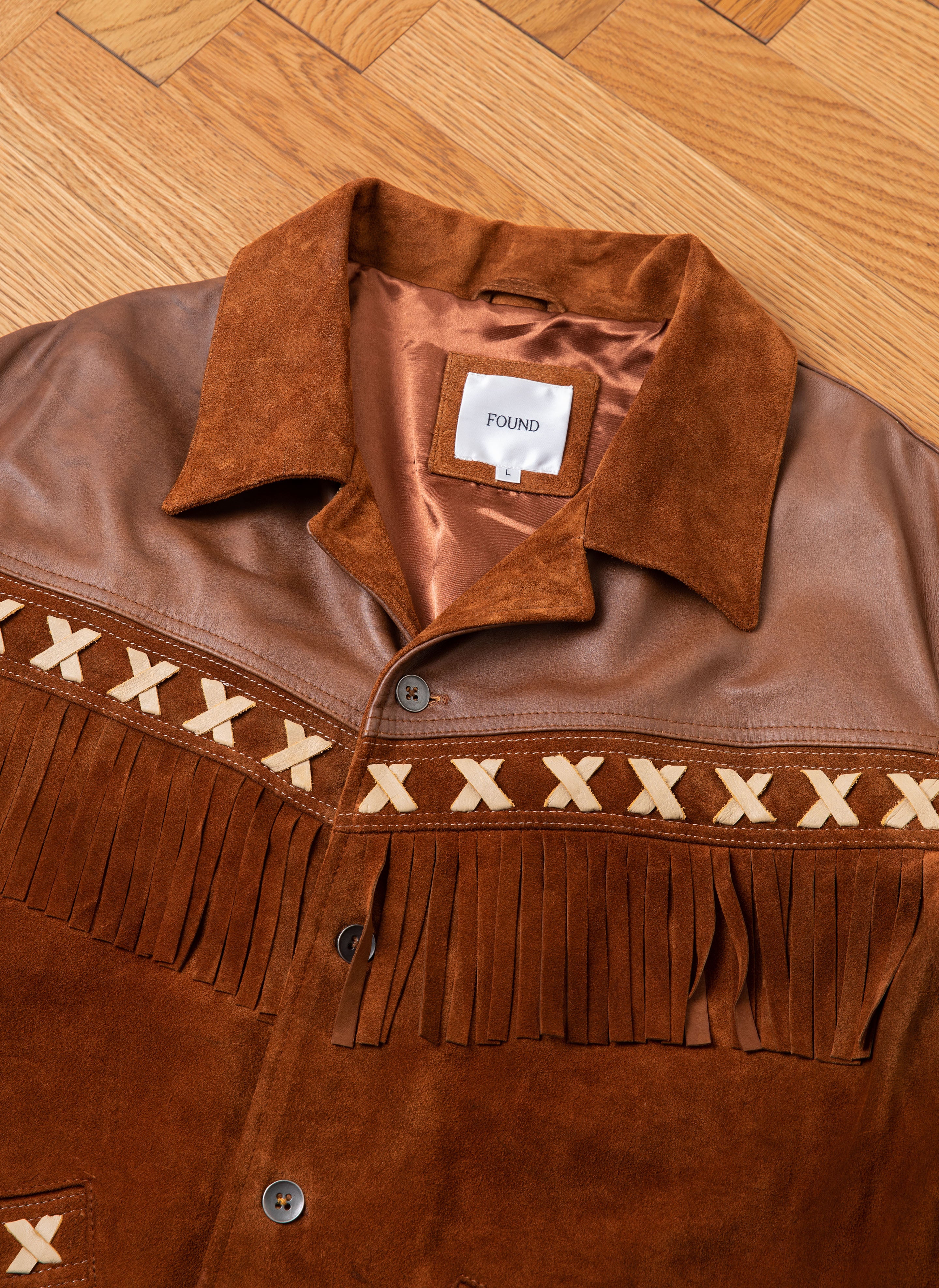 The Western Fringe Suede Leather Jacket by FOUND is made from genuine leather and ethically sourced materials. It features intricate X detailing on the chest, bold fringe accents, and a label inside that reads "FOUND," showcased beautifully on a wooden floor.