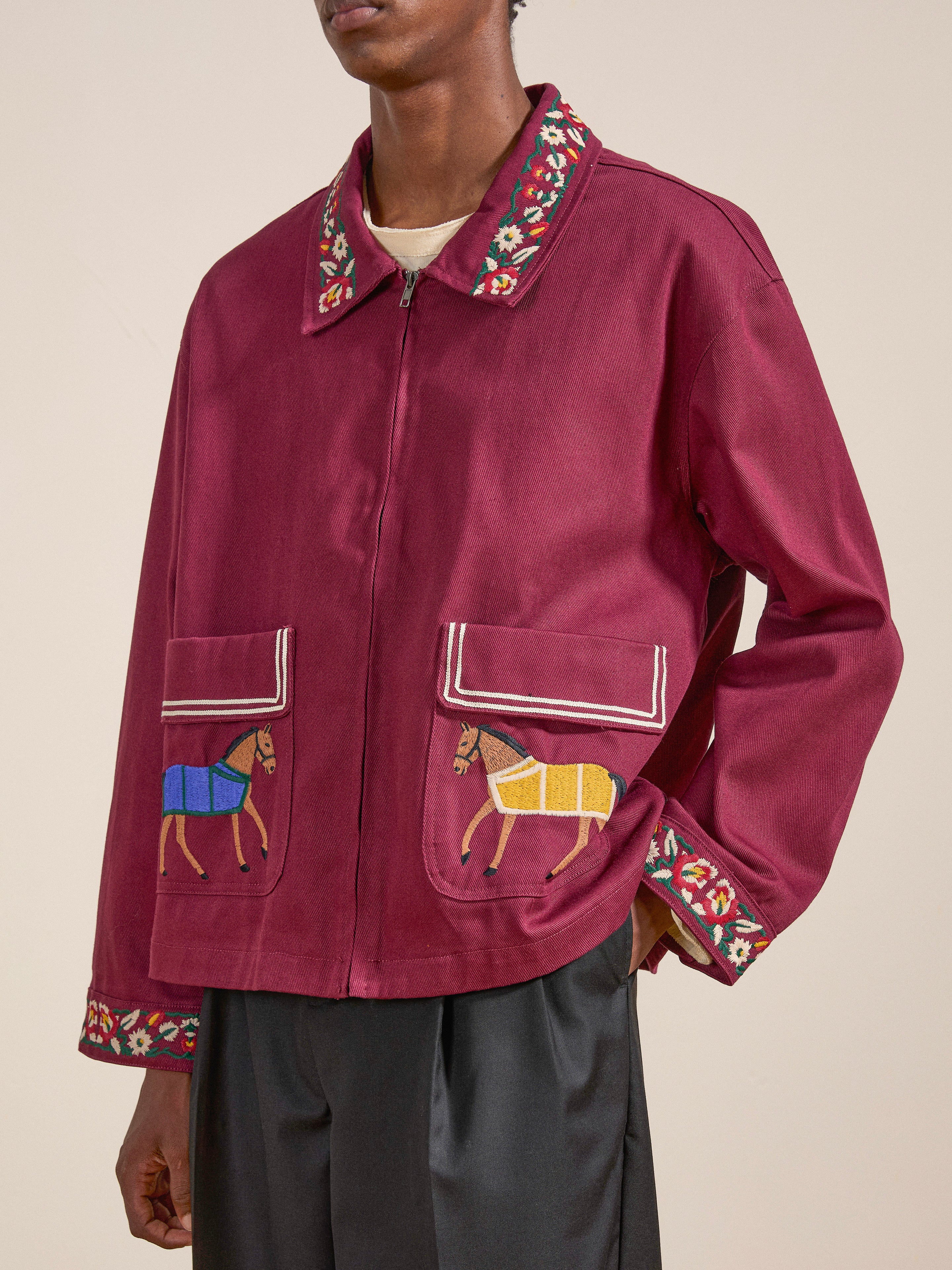 A person wears the Oxblood Horse Equine Jacket by FOUND, featuring intricate embroidery with horse designs on the front pockets and floral patterns on the collar and cuffs, reminiscent of 70’s jockey uniforms.