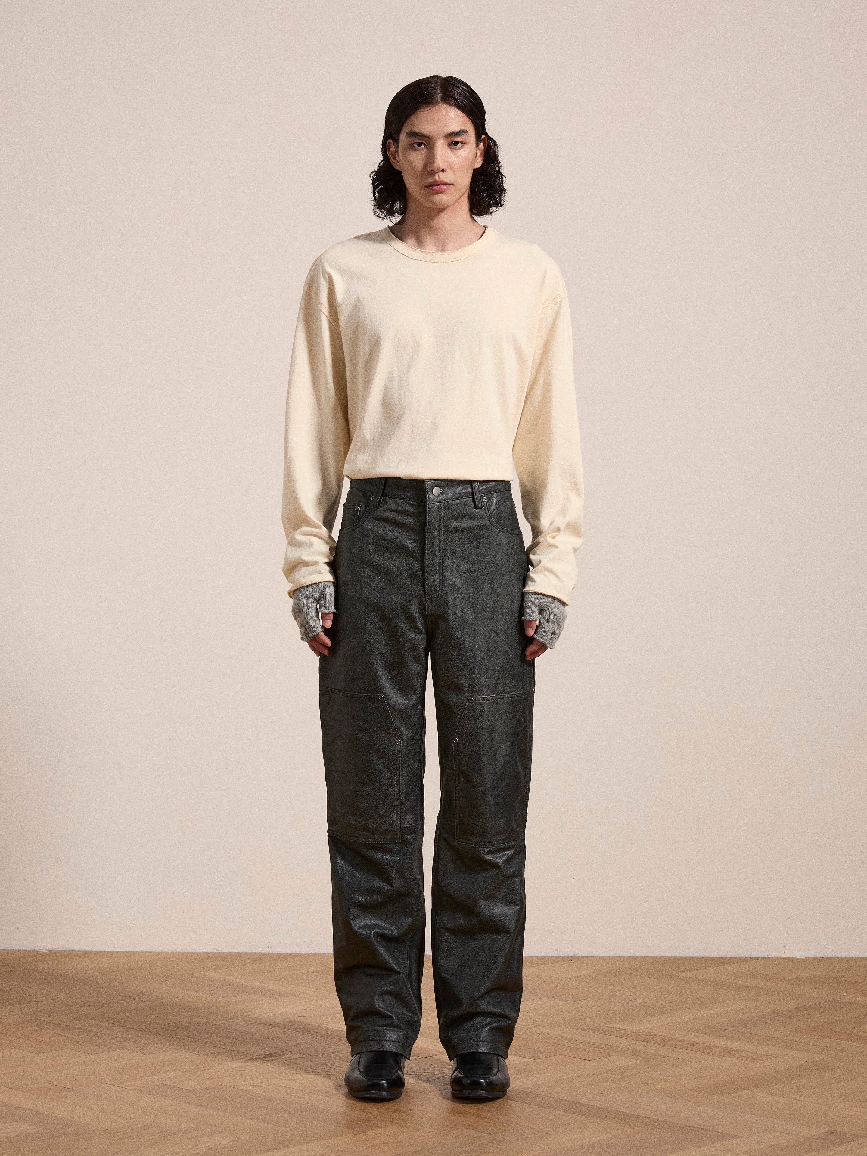 Person standing indoors against a plain background, wearing a long-sleeved, off-white shirt and dark, loose-fitting Distressed Genuine Leather Carpenter Pants from the brand FOUND, made from ethically sourced materials.