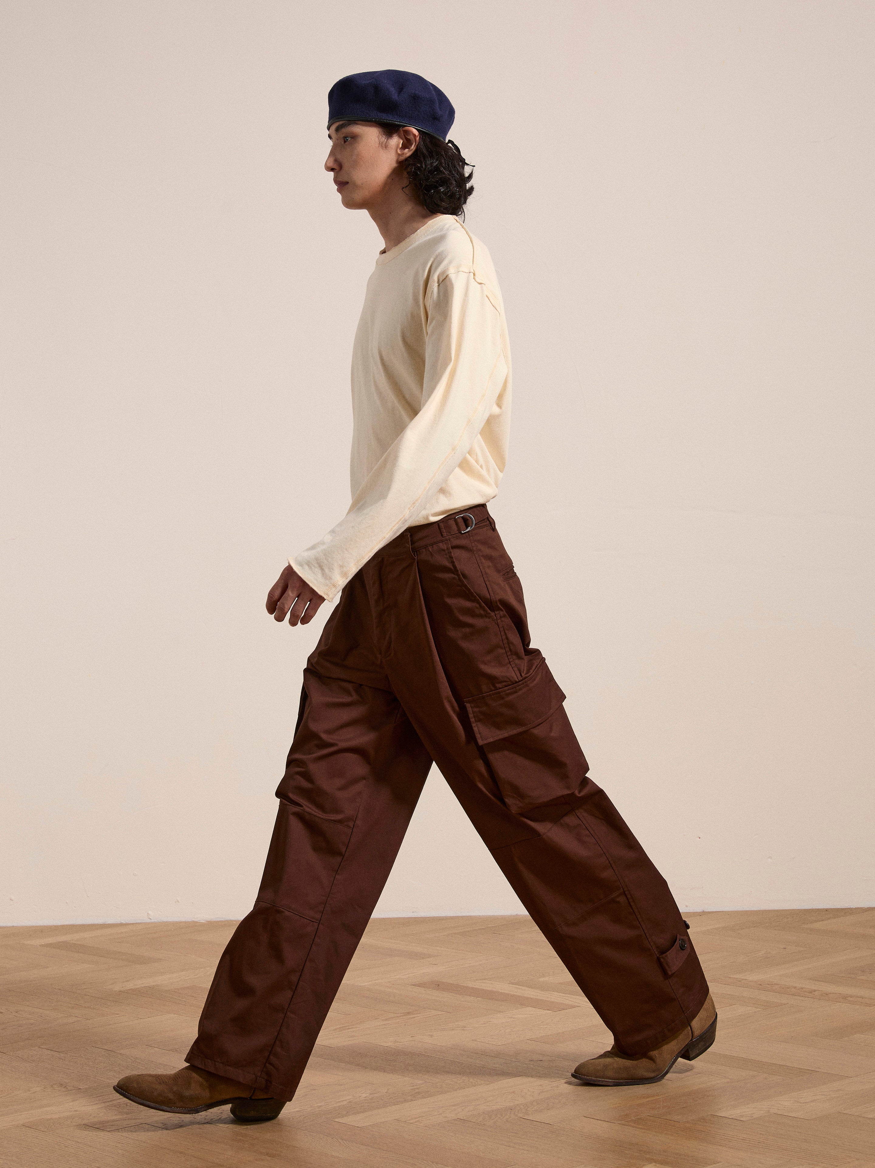 Person walking side profile indoors, wearing a blue beret, a cream long-sleeve shirt, FOUND Pleated Pocket Trousers, and brown shoes.